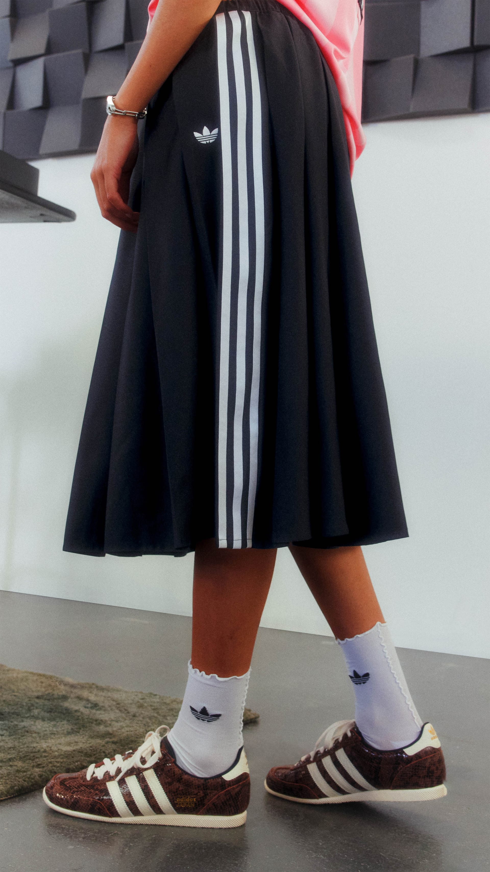 A person wearing a black skirt with white stripes, white socks, and brown sneakers stands in front of a gray and black textured background.