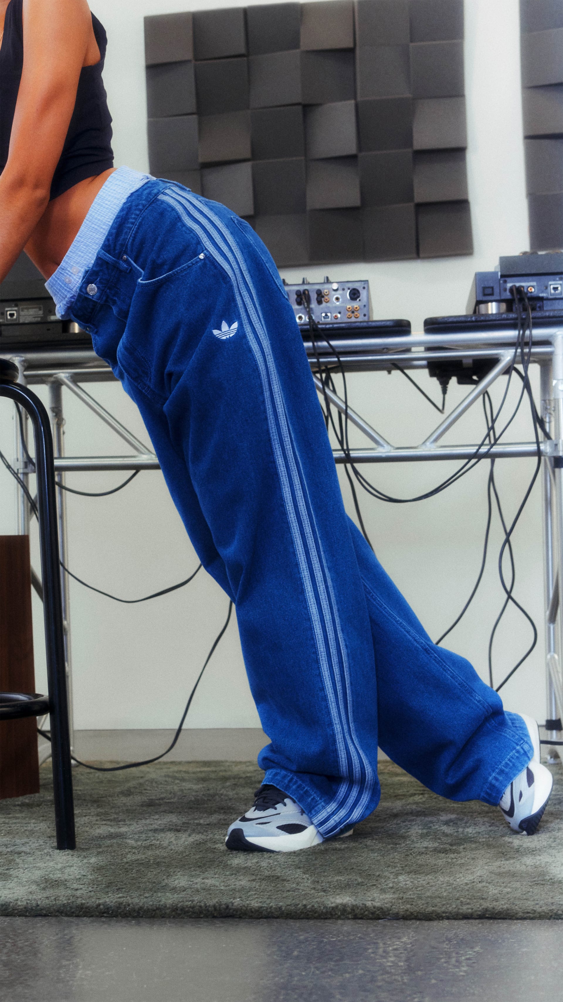 A person wearing blue Adidas tracksuit pants and white sneakers is sitting in front of a wall with a textured, geometric pattern.