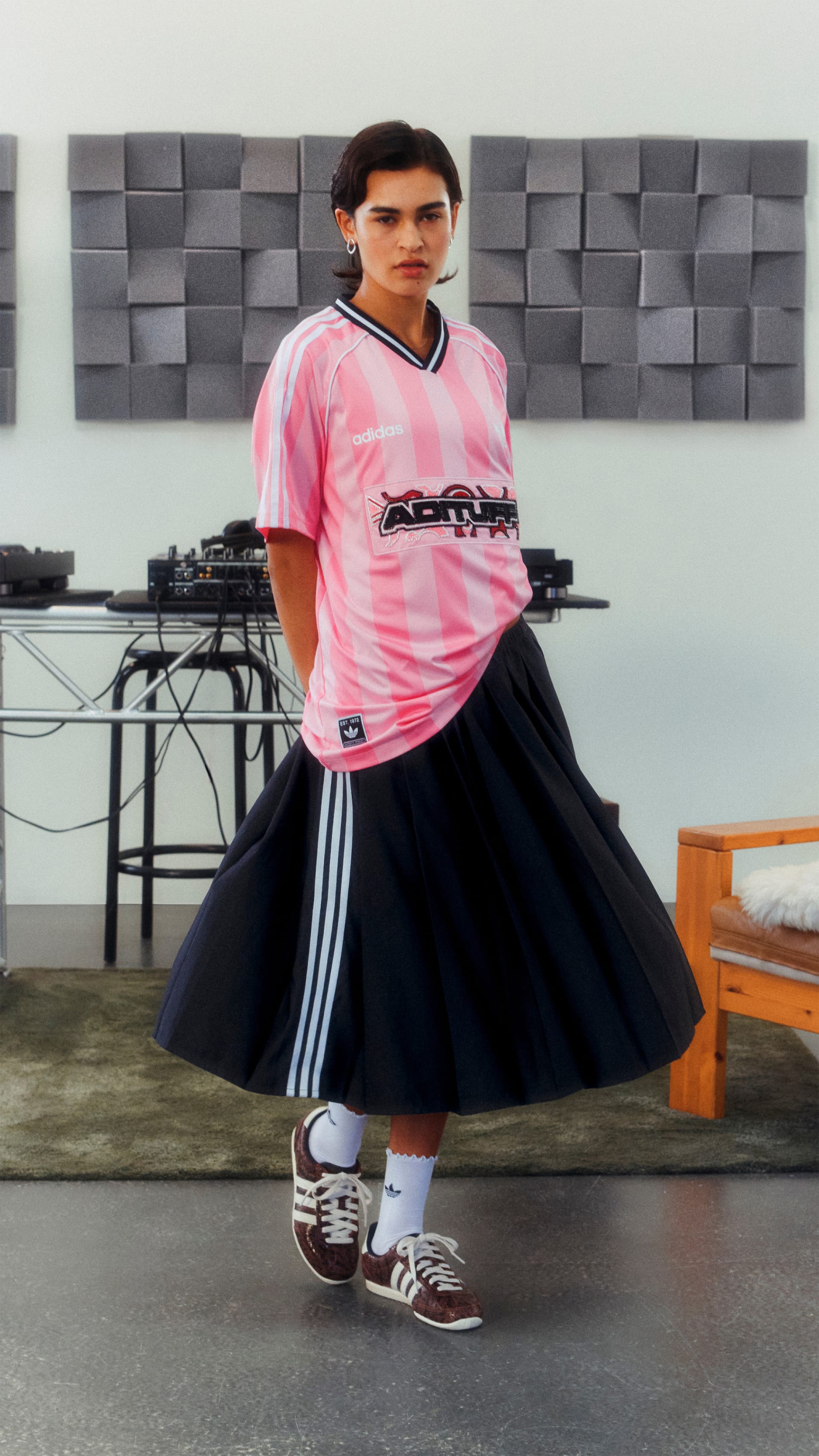 A person wearing a pink t-shirt and a black skirt stands in front of a gray textured wall, with various audio equipment visible in the background.