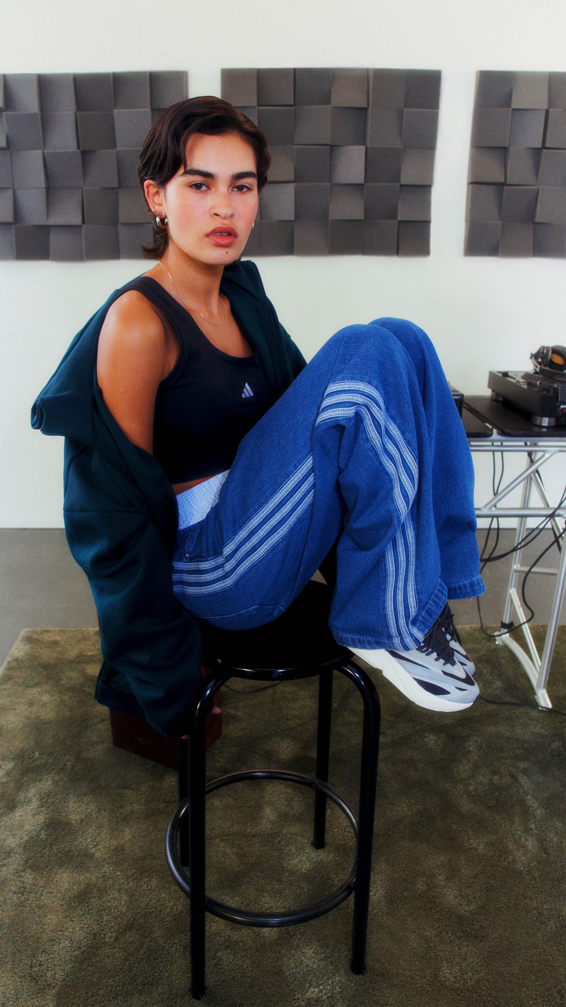 A person wearing a black top and blue pants is sitting on a stool in front of a wall with a textured pattern.