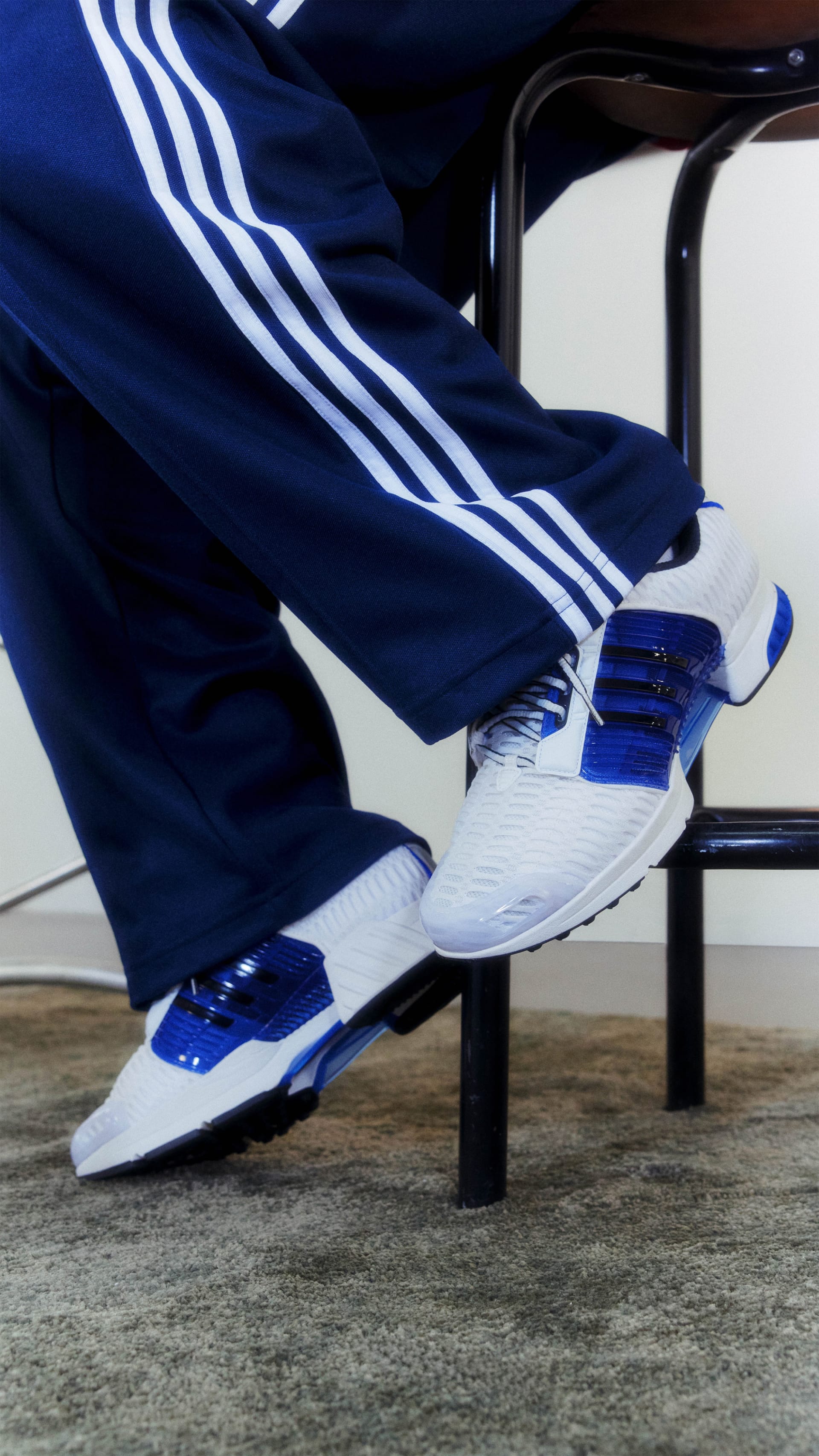 A pair of white and blue athletic shoes with three stripes on the side, worn by a person sitting on a chair against a dark background.