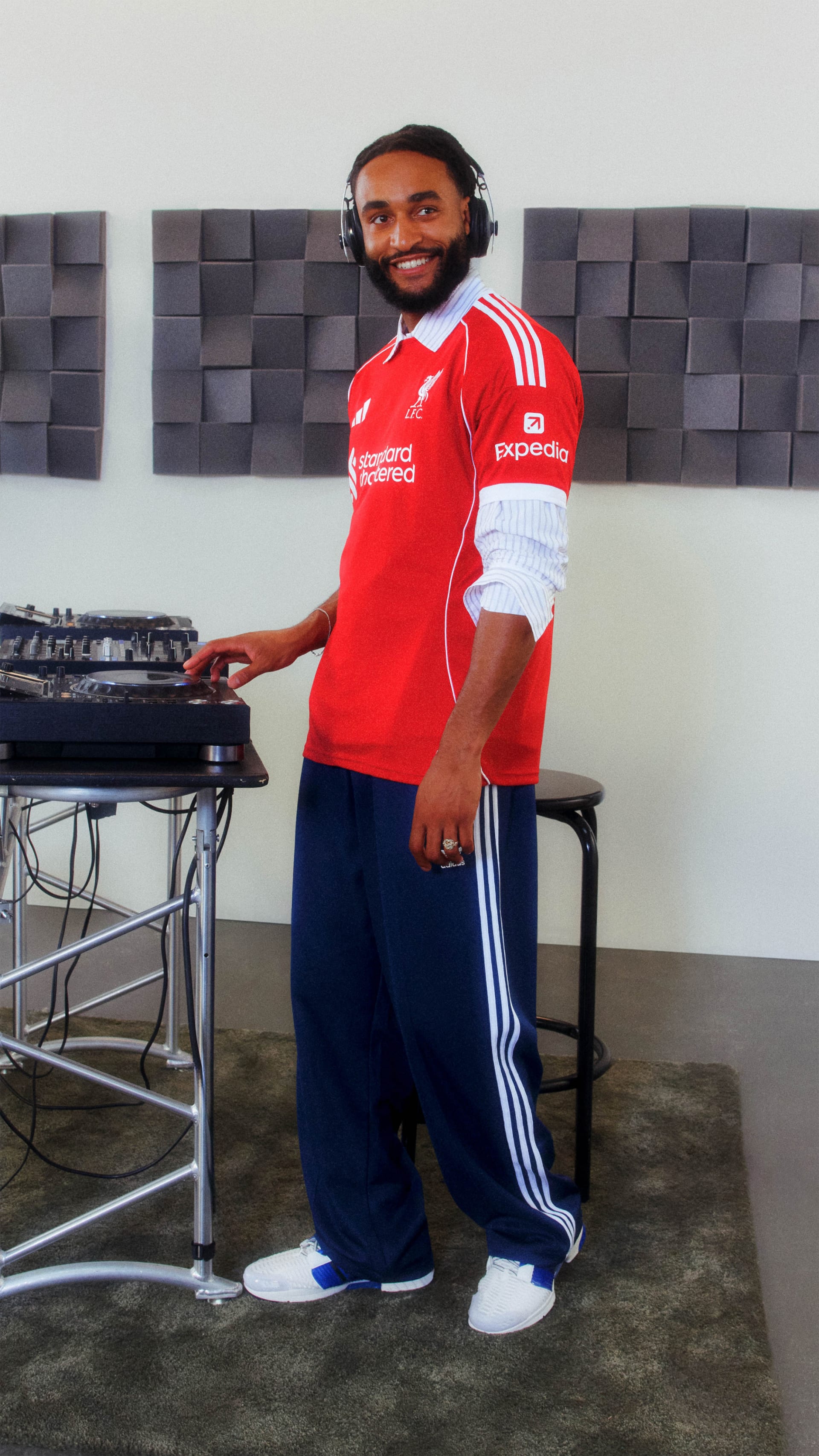 A smiling man wearing a red sports jersey and headphones stands in front of a wall with geometric patterns, surrounded by audio equipment.