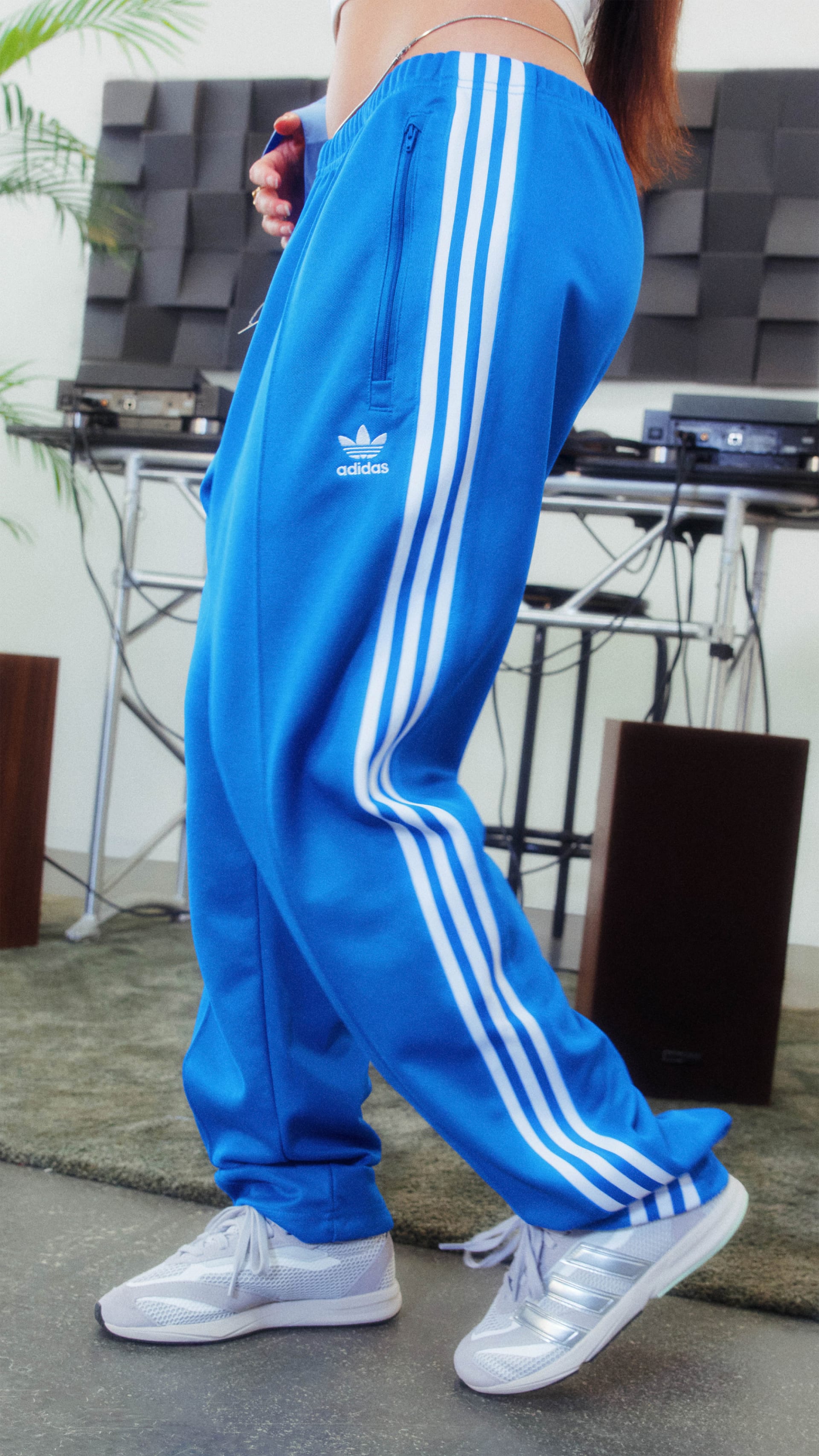 The image shows a person wearing blue Adidas tracksuit pants with white stripes, standing in a room with a television and other electronic equipment in the background.