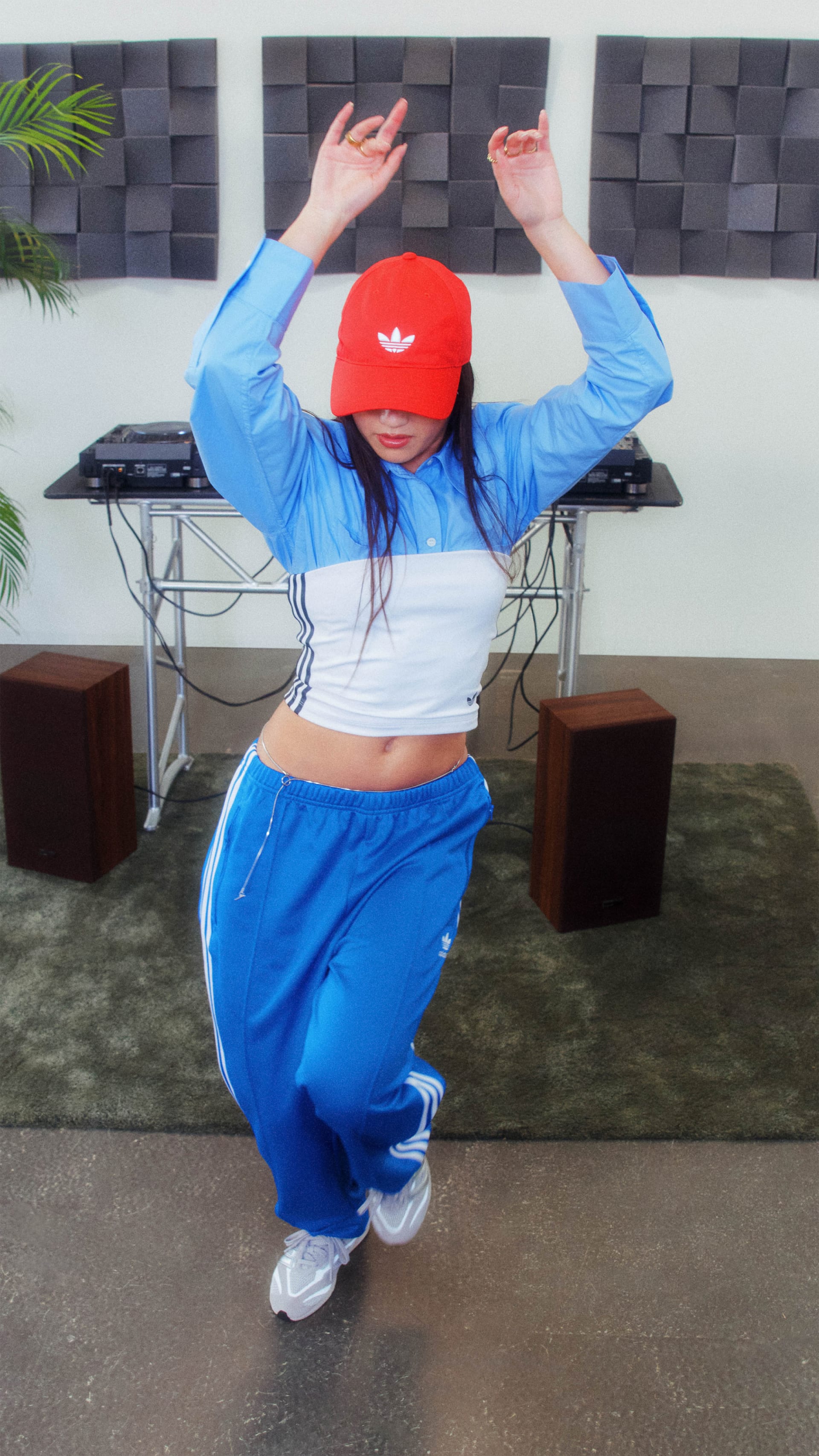 A person wearing a red cap, blue crop top, and matching blue pants is dancing with their arms raised in a room with a textured gray wall and potted plant in the background.