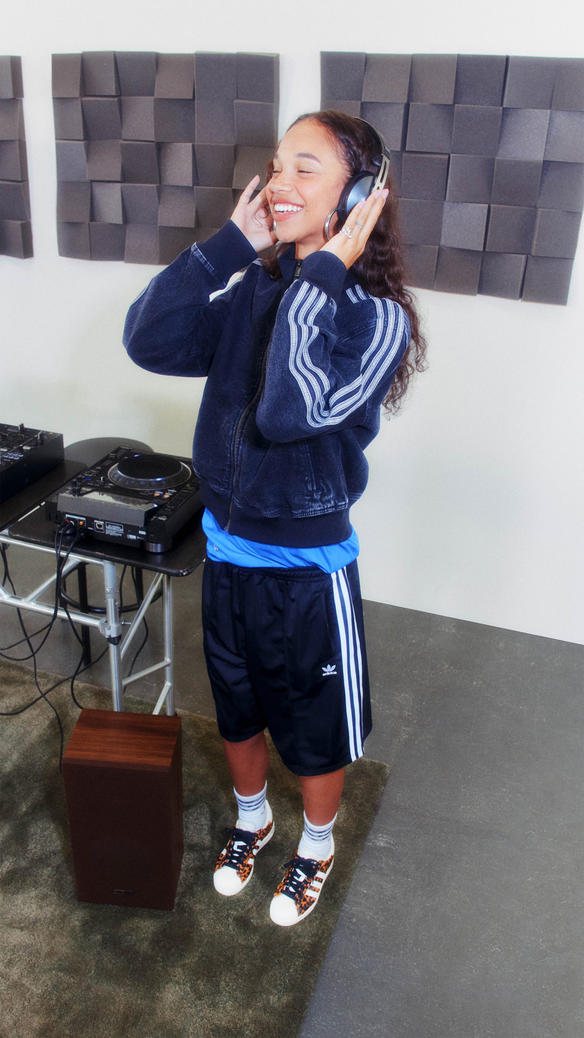 A young woman wearing a navy blue tracksuit and headphones stands in a room with gray textured walls, appearing to be listening to music or engaged in an audio activity.