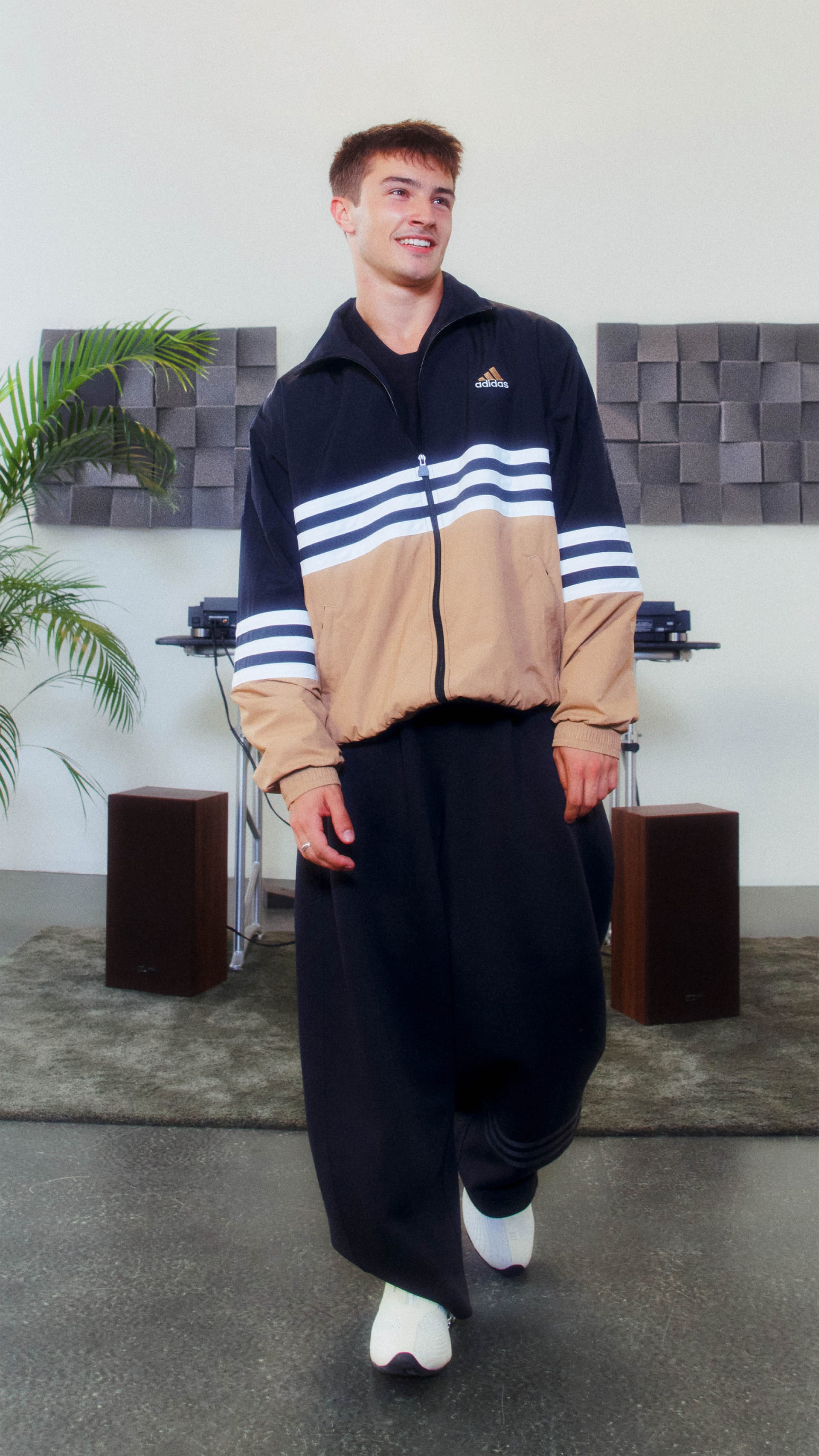 A young man wearing a black and beige striped jacket stands in a room with a modern, minimalist decor featuring gray cubes and a potted plant in the background.
