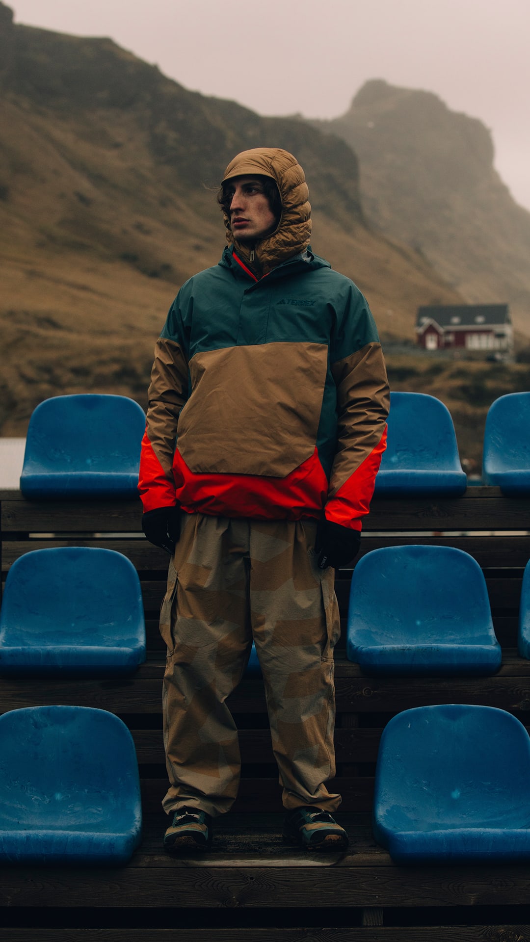 A person wearing warm outdoor clothing stands on a bench in a mountainous landscape, with a vehicle visible in the background.