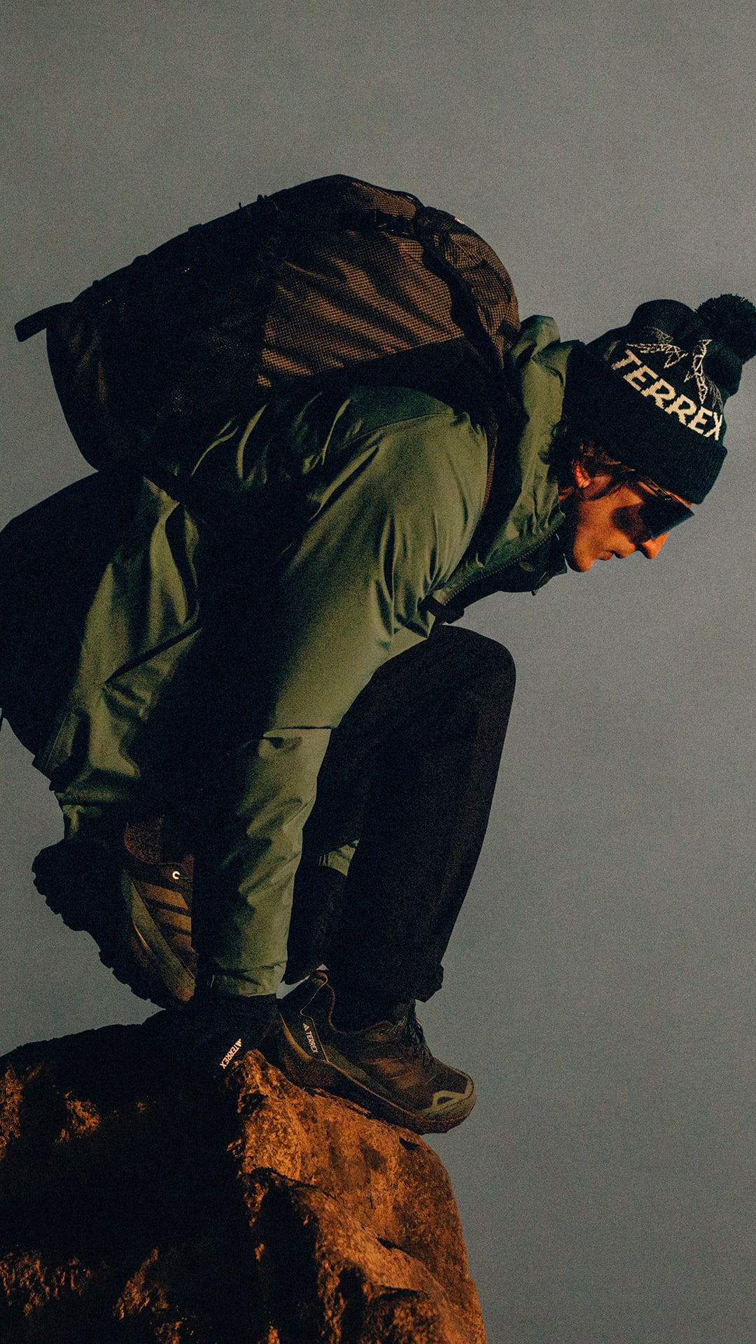 A person wearing a dark jacket and hat is crouched on a rocky surface against a dark background.