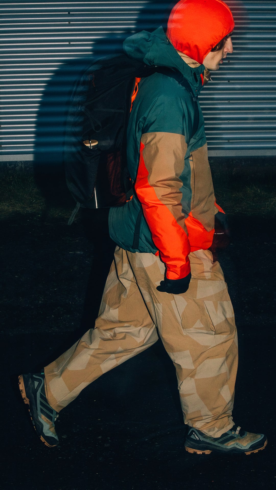 A person wearing a bright red hat, green jacket, and beige pants is walking in front of a metal shutter backdrop.