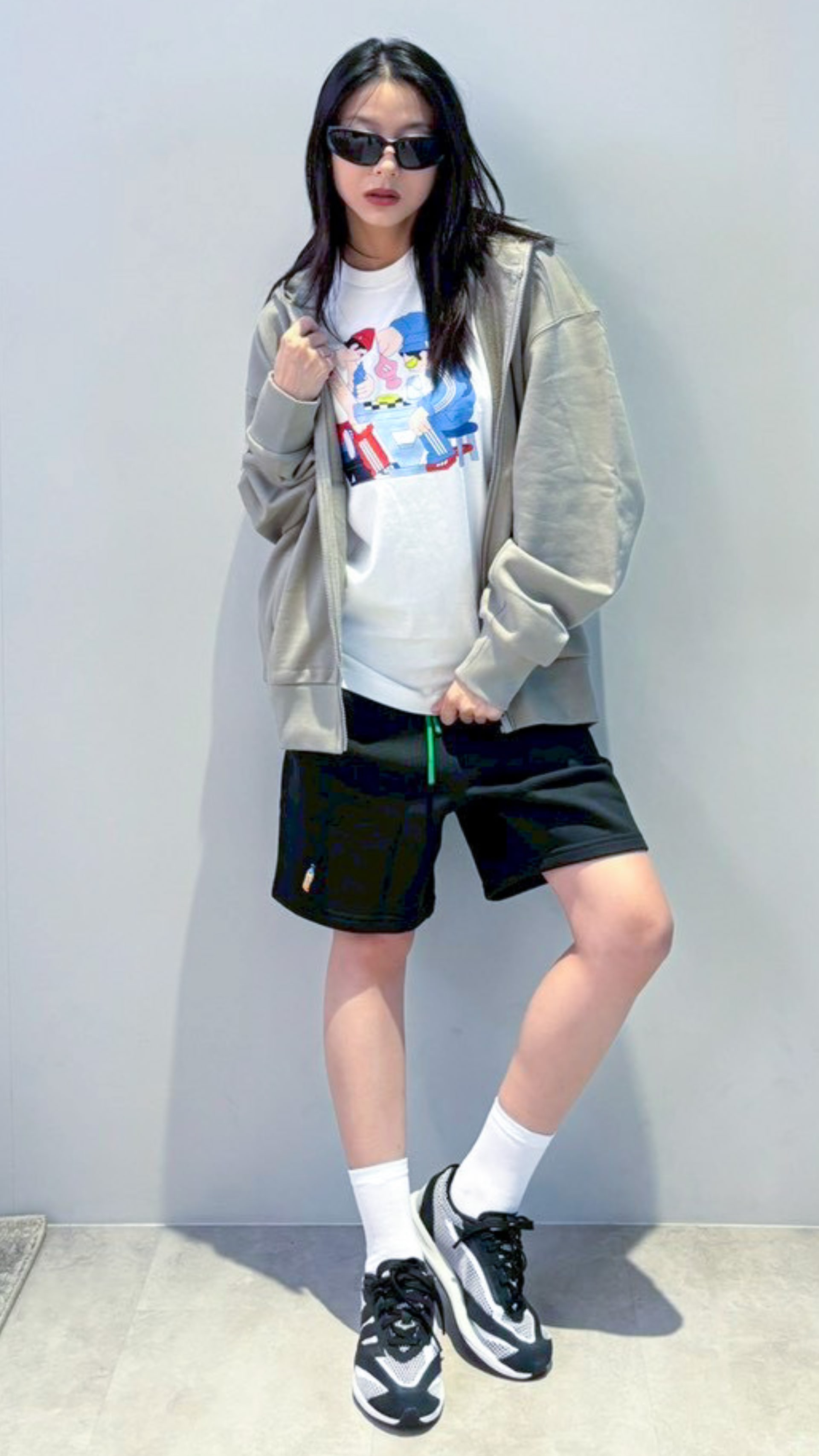 The image depicts a young woman wearing a light-colored jacket, a graphic t-shirt, and black shorts. She is standing against a plain white background and appears to be posing for the camera.