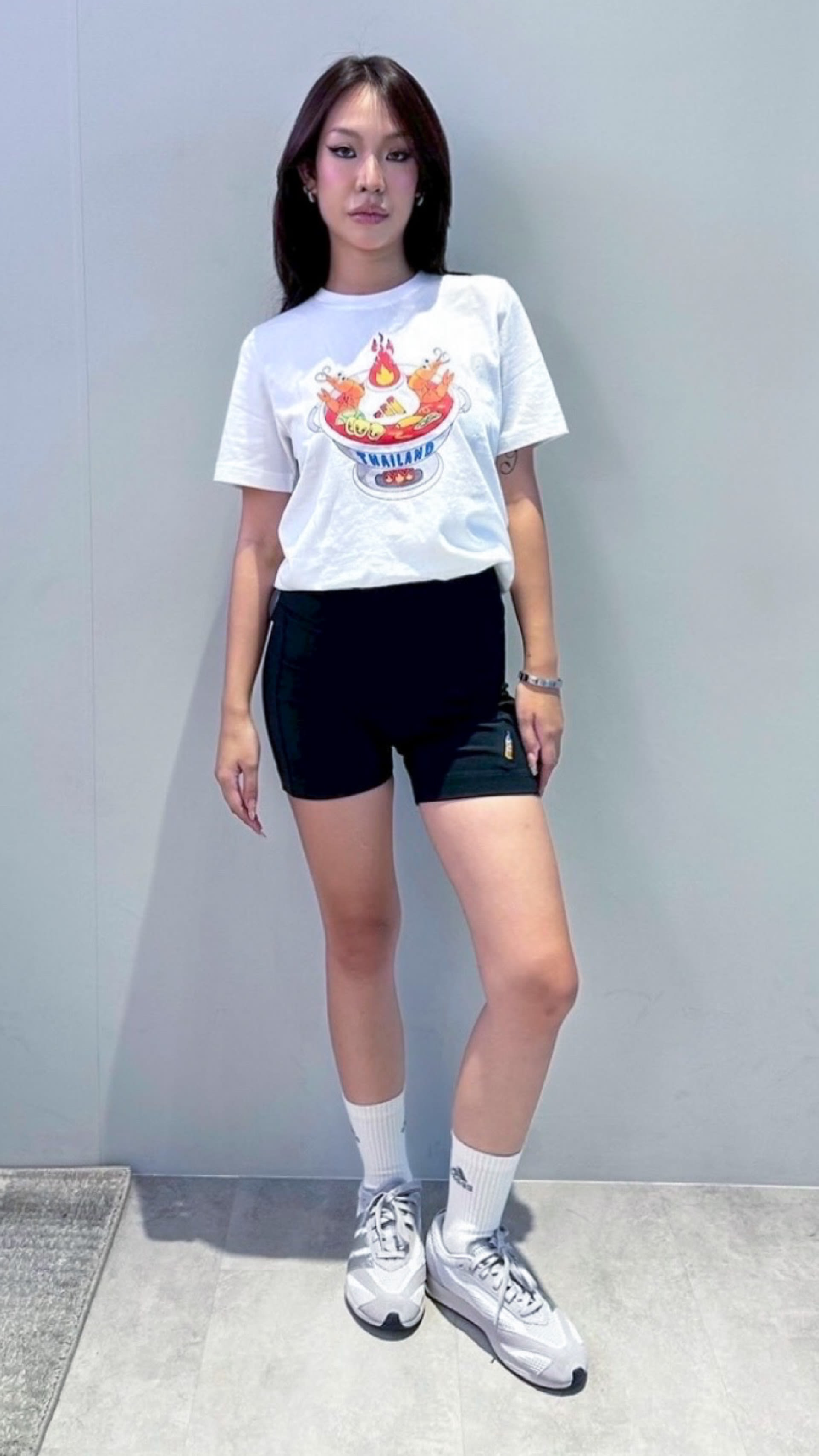 A young woman wearing a white t-shirt with a graphic design, black shorts, and white sneakers stands in front of a plain gray background.