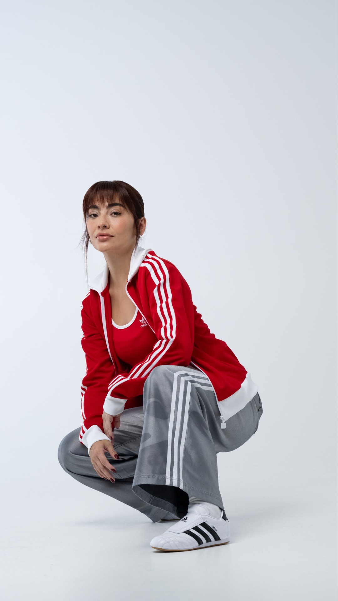A young woman wearing a red jacket and gray pants is sitting on the floor against a plain white background.