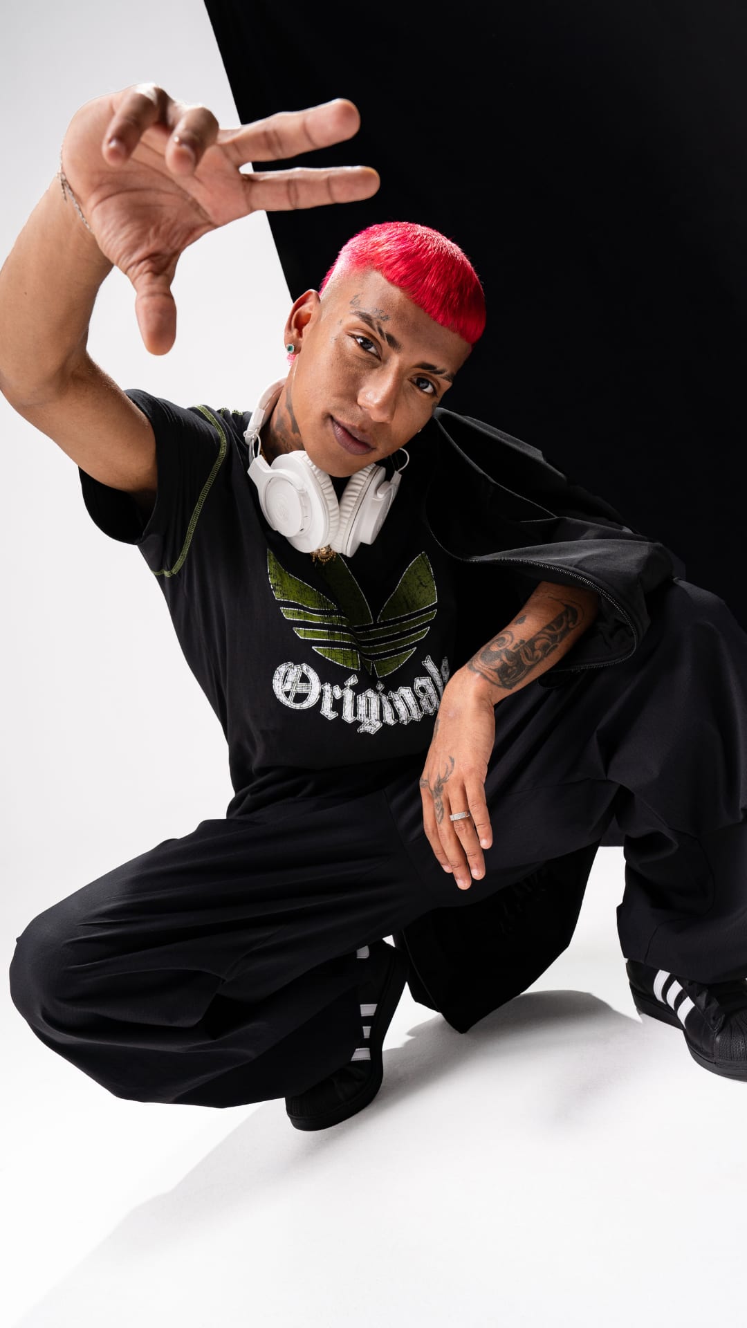 A person with red hair and tattoos, wearing a black shirt with a logo, is sitting on the floor and making a hand gesture against a plain white background.