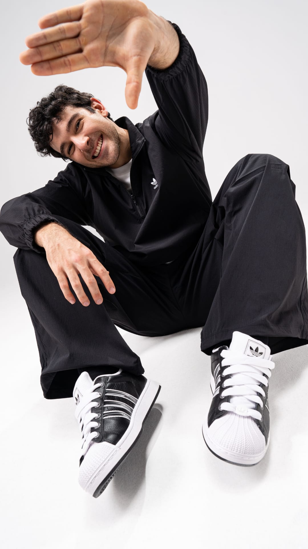 A person wearing a black outfit and white sneakers is sitting on the floor, with their hand raised in a gesture.