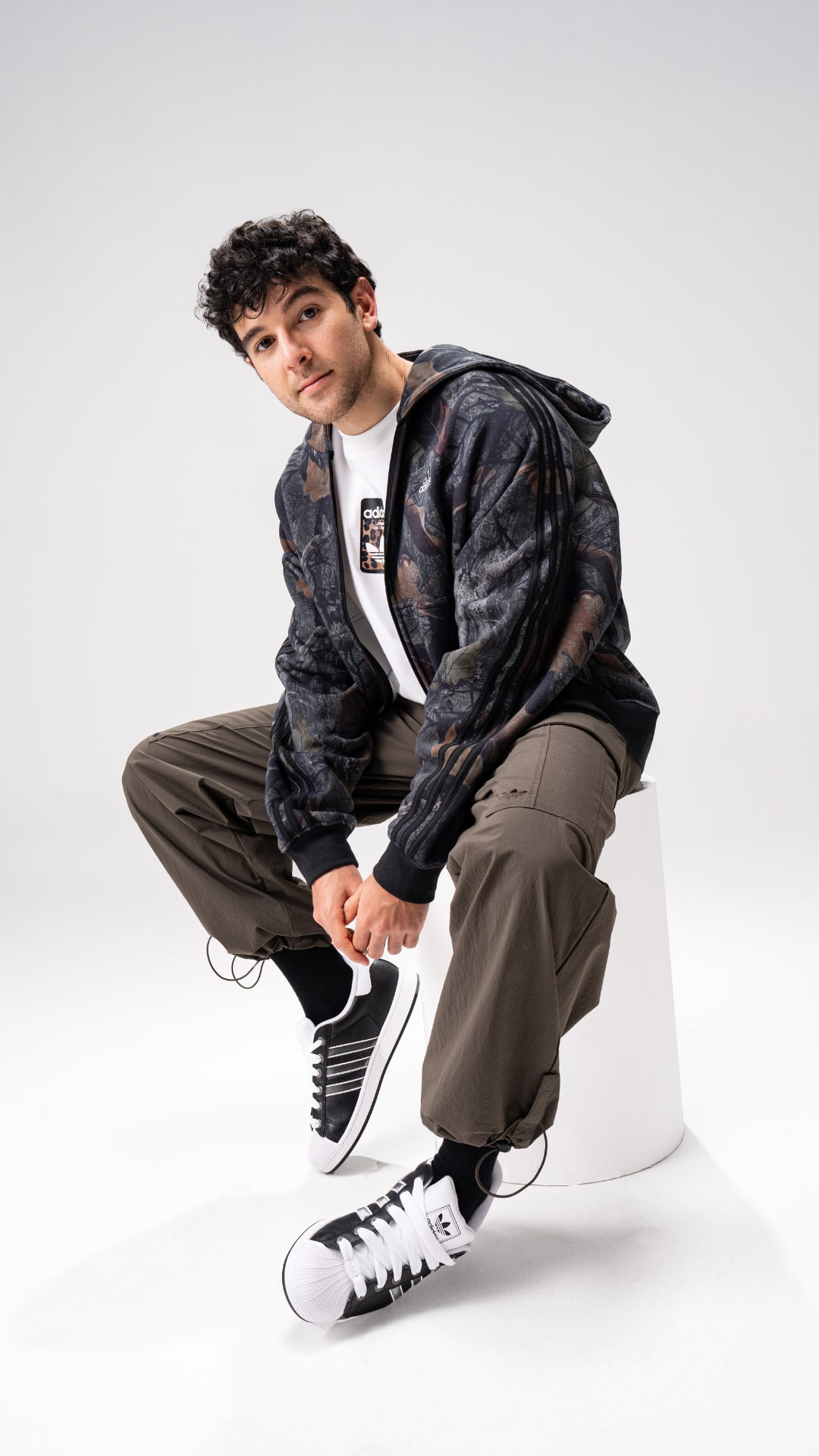 A young man with curly hair wearing a dark jacket, white t-shirt, and khaki pants is sitting on a white cube against a plain background.