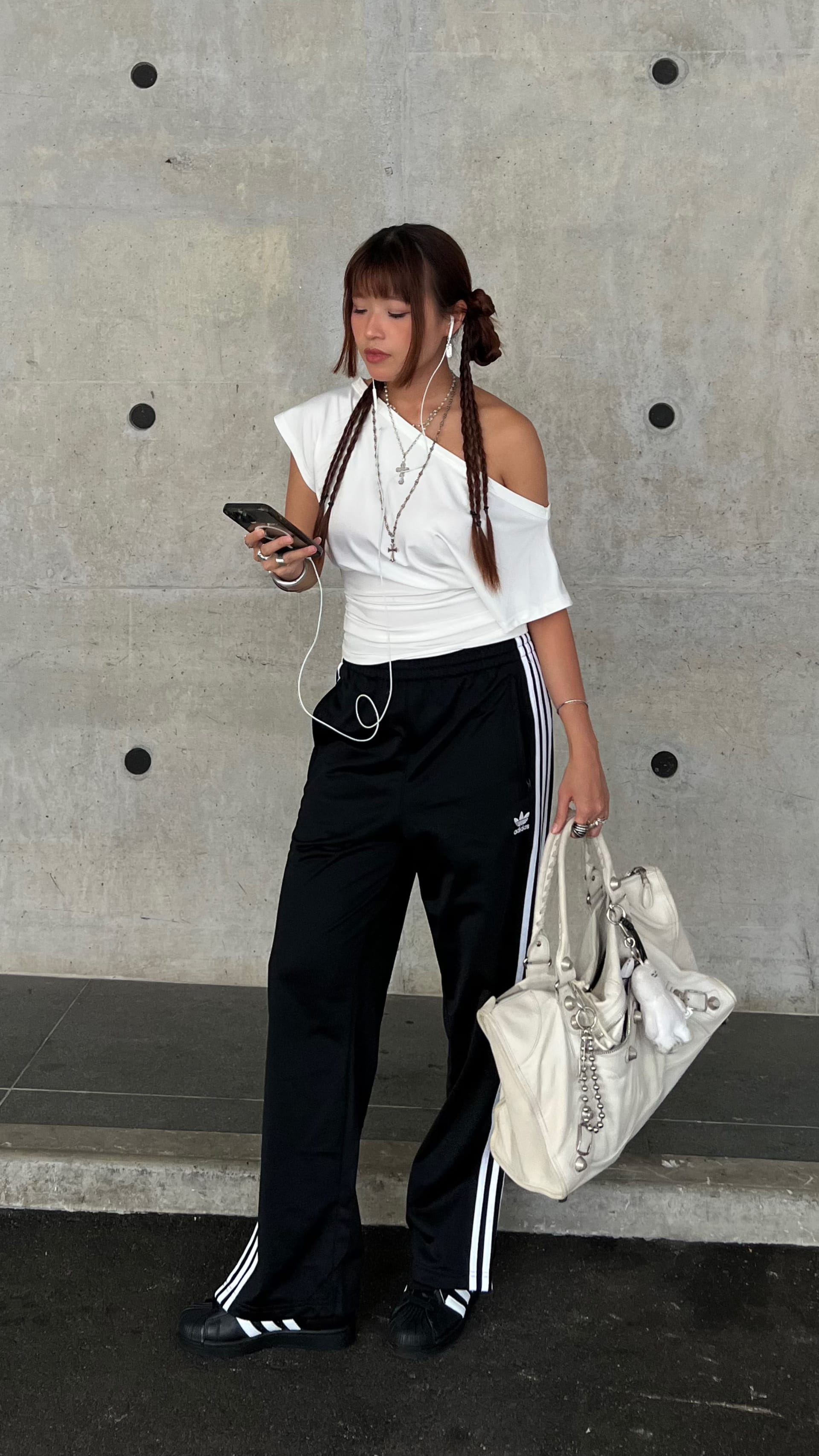 A young woman with braided hair stands in front of a concrete wall, holding a mobile phone and a bag, wearing a white off-the-shoulder top and black pants with stripes.