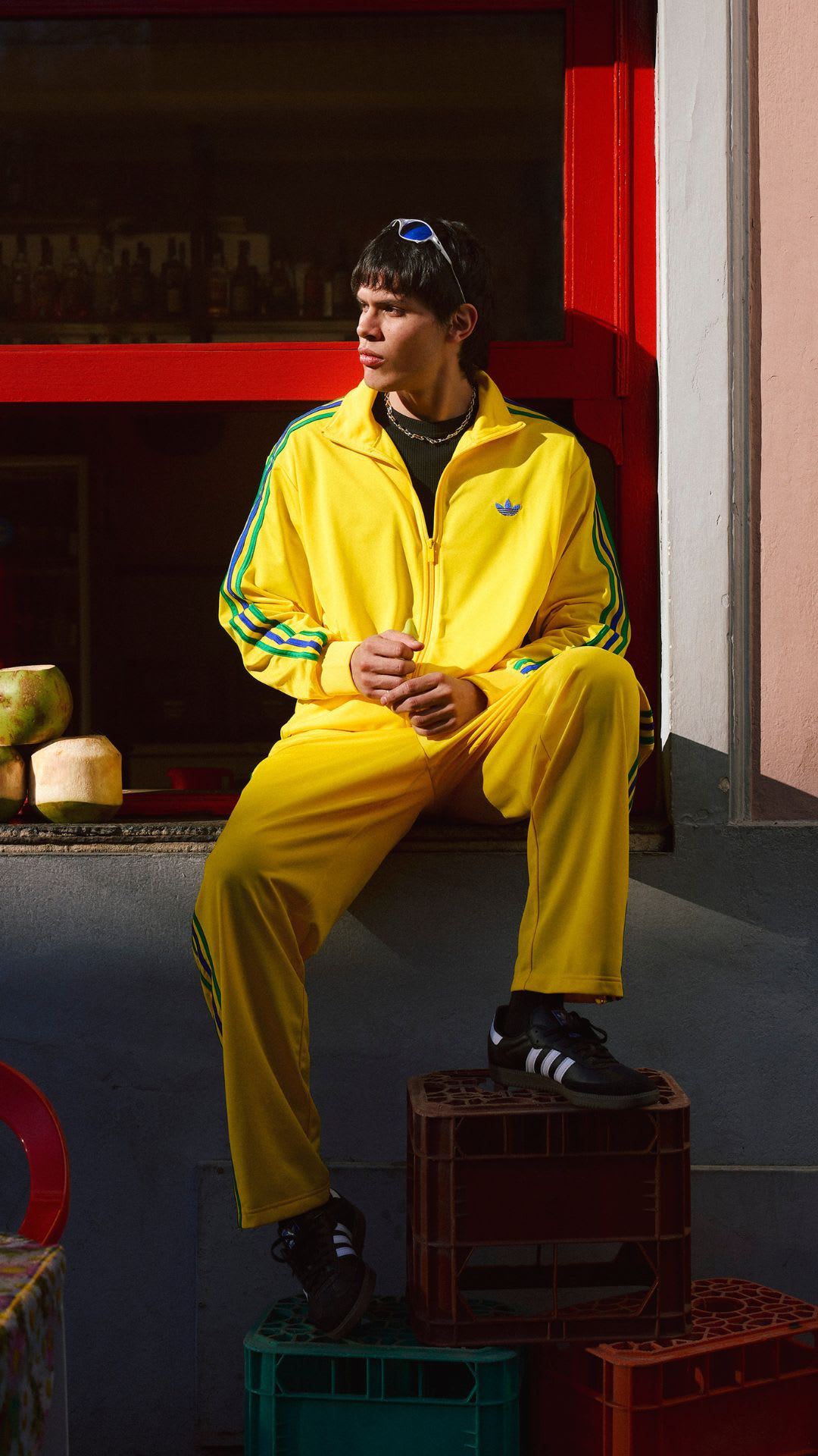 A person wearing a bright yellow tracksuit is sitting on a wooden crate in a room with red walls and shelves in the background.