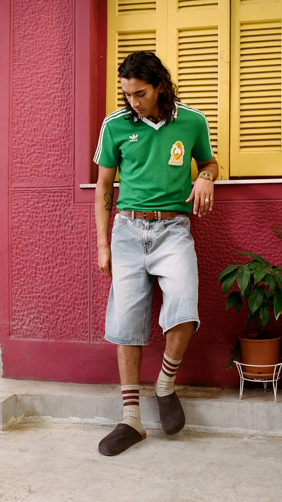 A person wearing a green jersey and light-colored shorts is standing in front of a vibrant pink wall, with potted plants visible in the background.