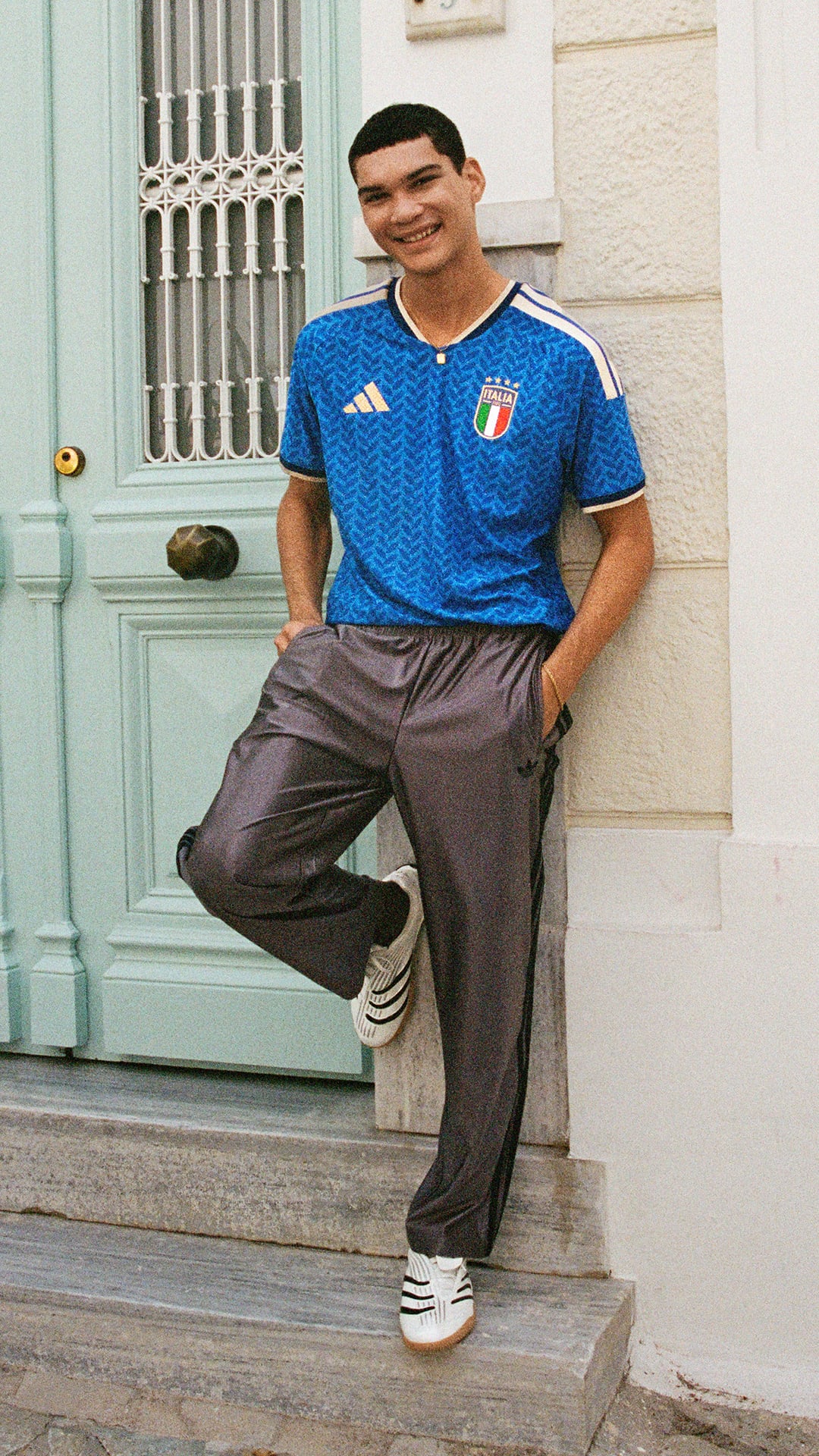 A young man wearing a blue sports jersey and loose-fitting pants is standing on the steps of a building with a green door in the background.