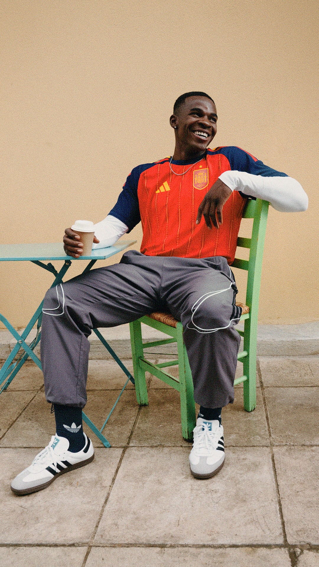 A man wearing an orange jersey and casual clothing is sitting on a colorful chair, holding a cup and appearing relaxed in an outdoor setting with a plain background.