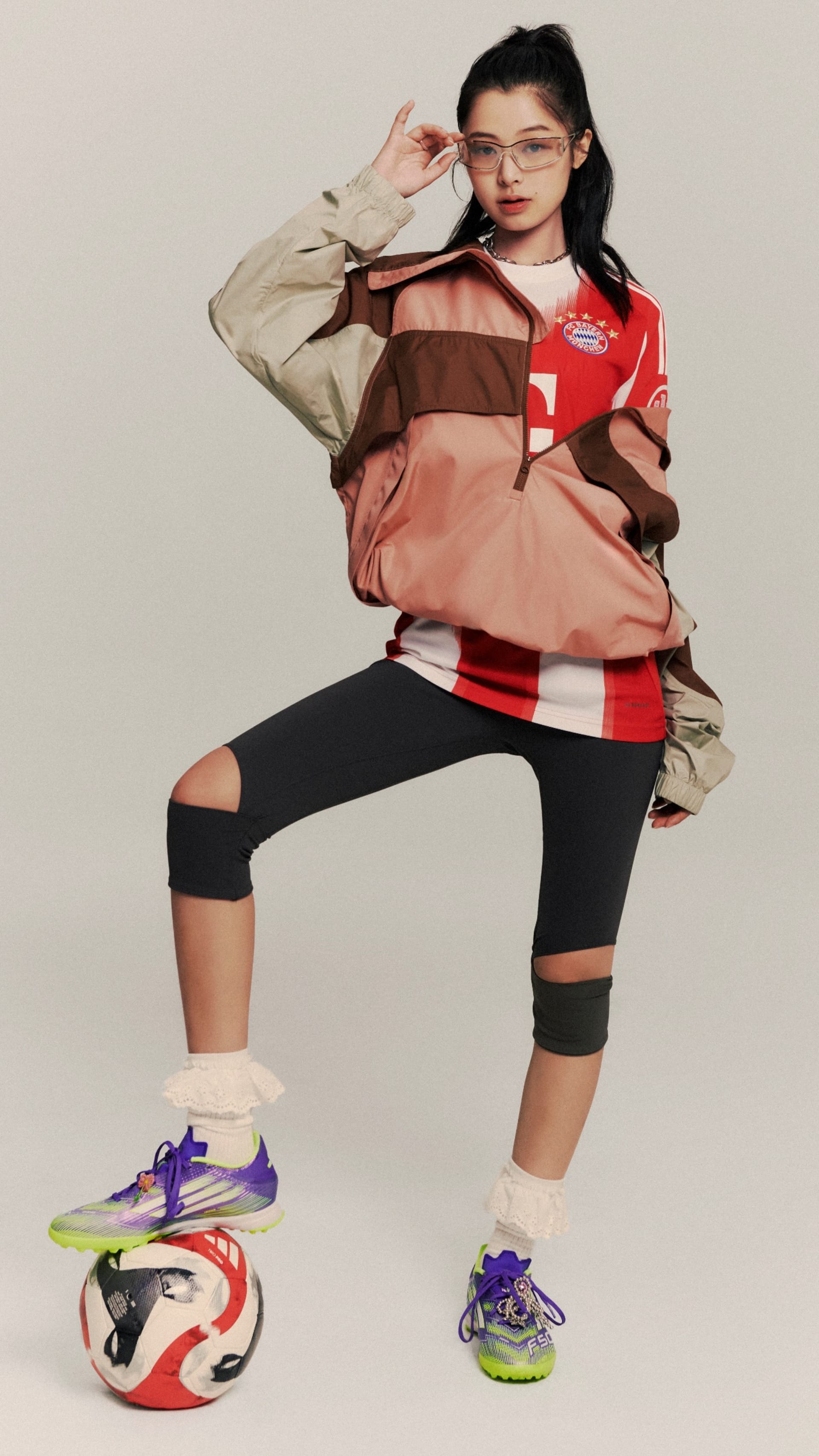 A young woman wearing a red and white athletic top, black leggings, and colorful sneakers is posing with a soccer ball on a plain background.