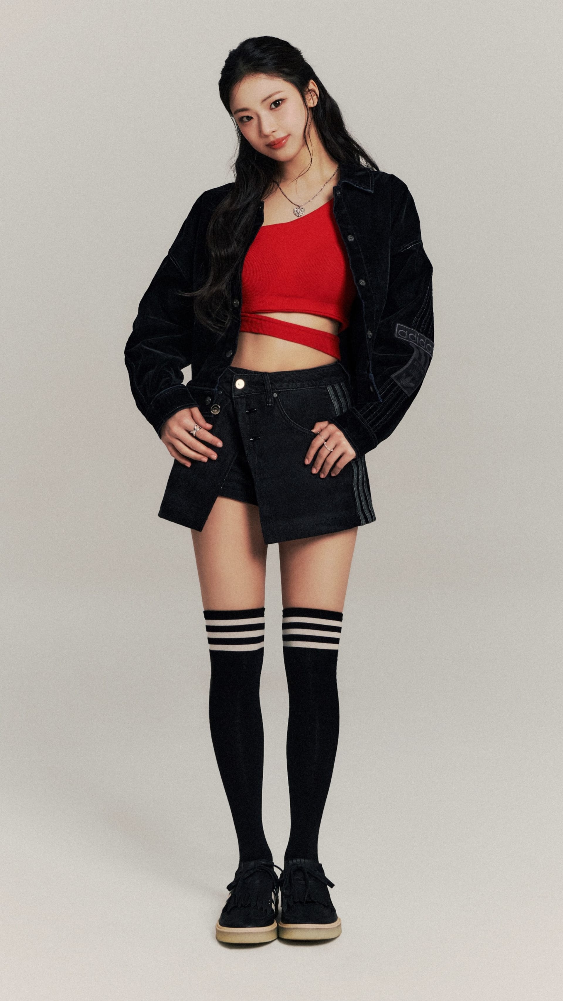 A young woman wearing a red crop top, black shorts, and knee-high socks stands against a plain background.