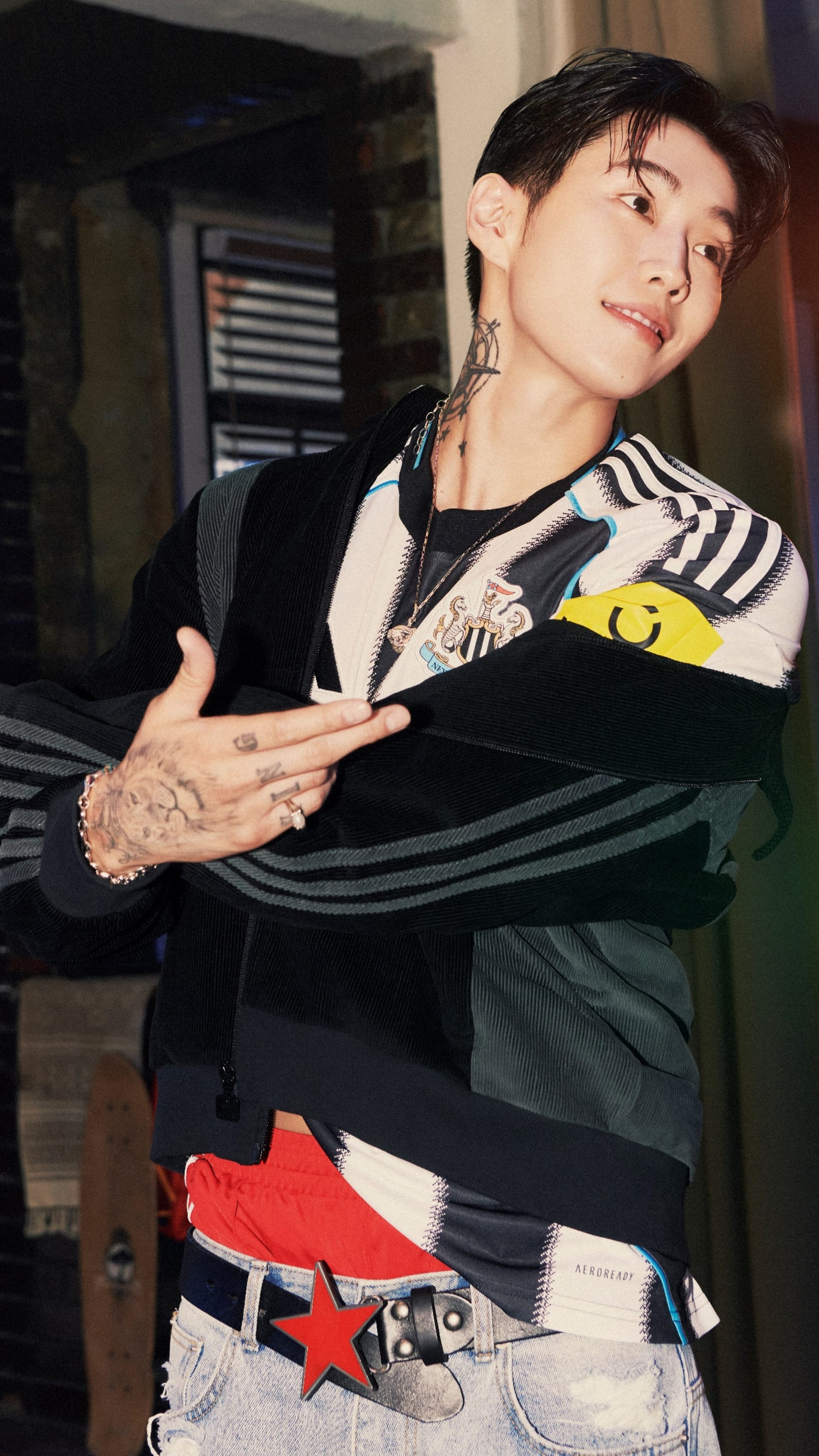 A person wearing a black jacket and a striped shirt is sitting in a room with various objects and furniture visible in the background.