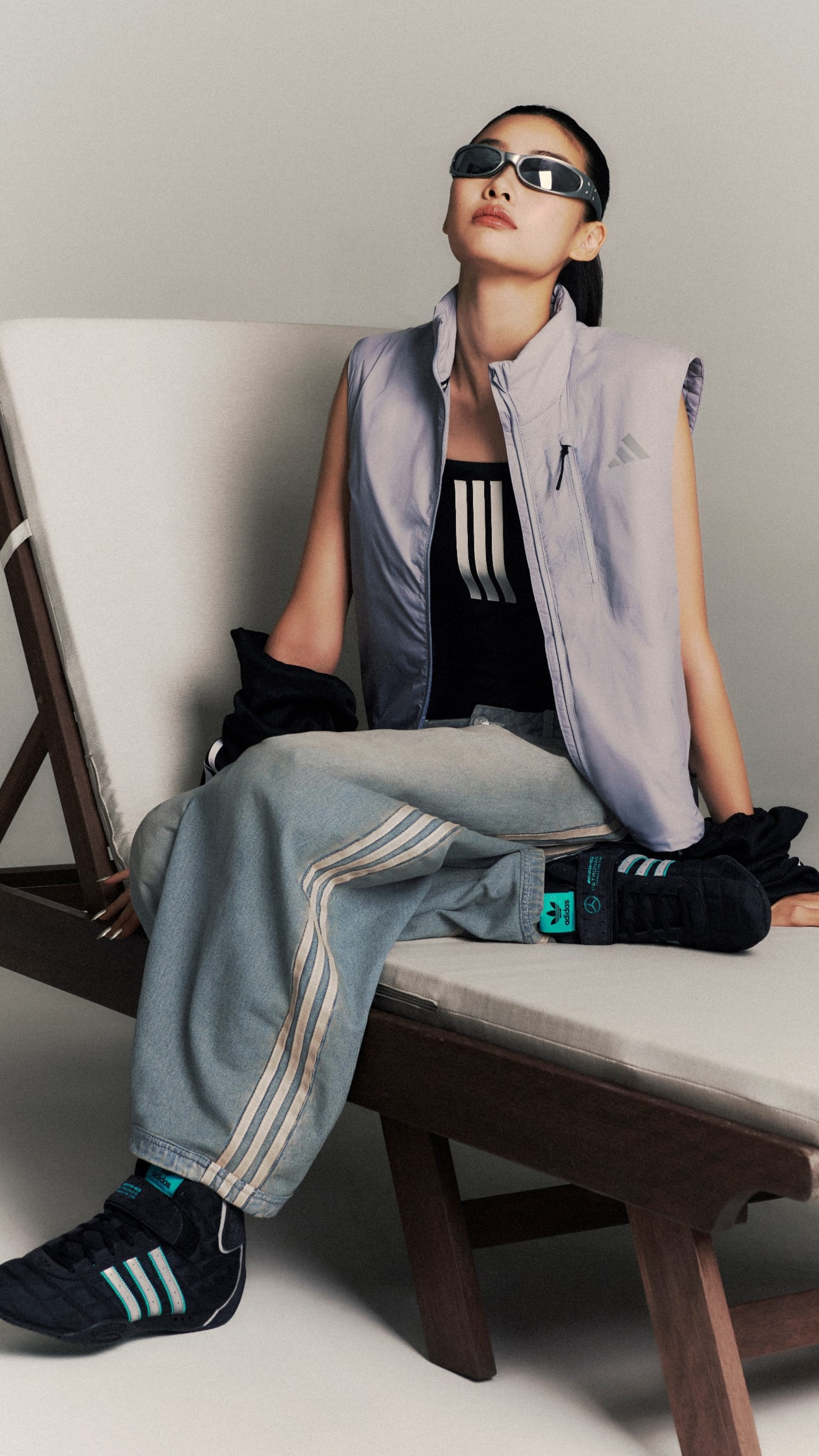 A person wearing a gray jacket, black top, and gray pants is sitting on a wooden bench, with a pair of sunglasses on their face.