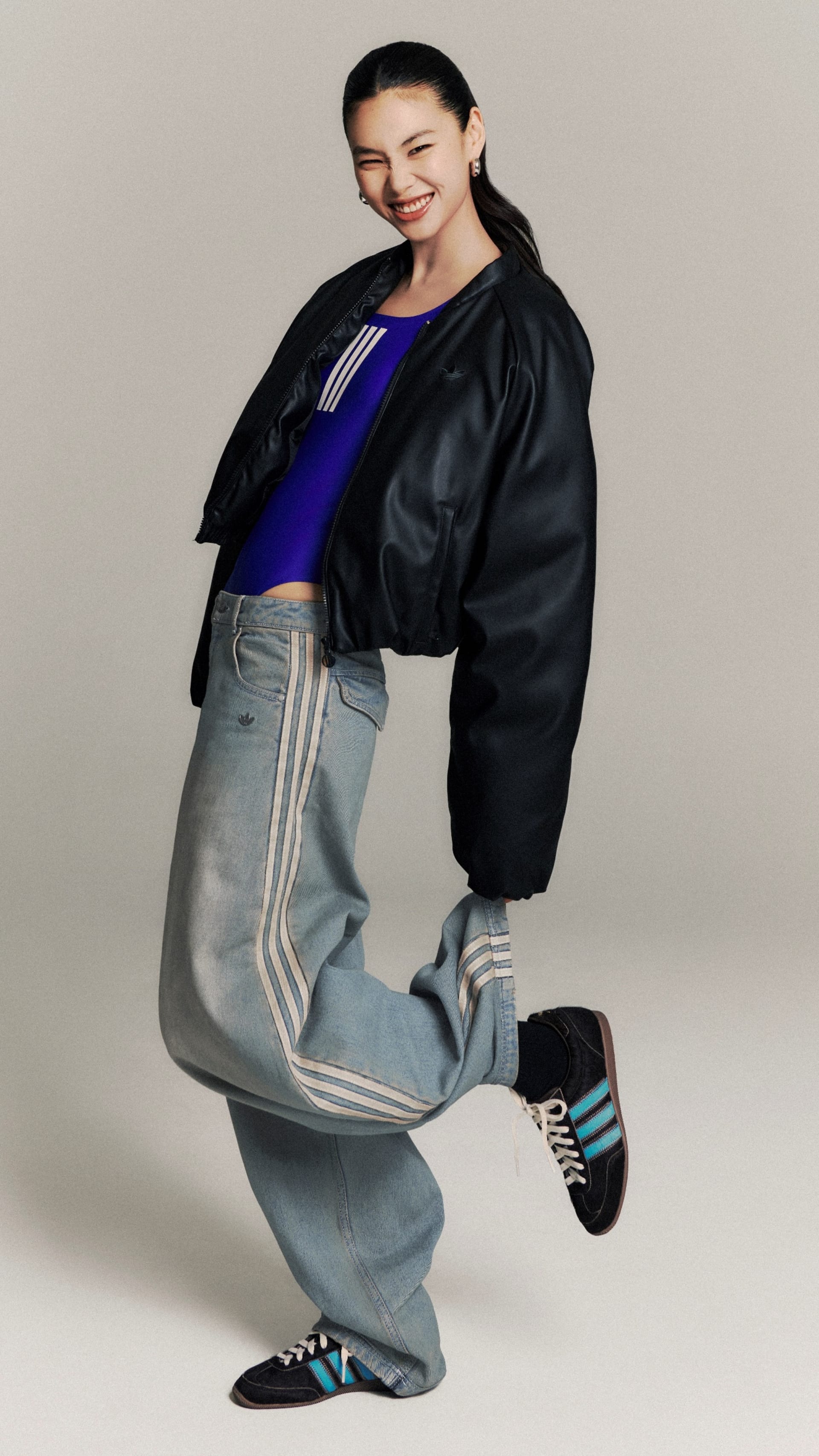 A smiling individual wearing a black leather jacket, a blue top, and gray pants with stripes, standing against a plain background.