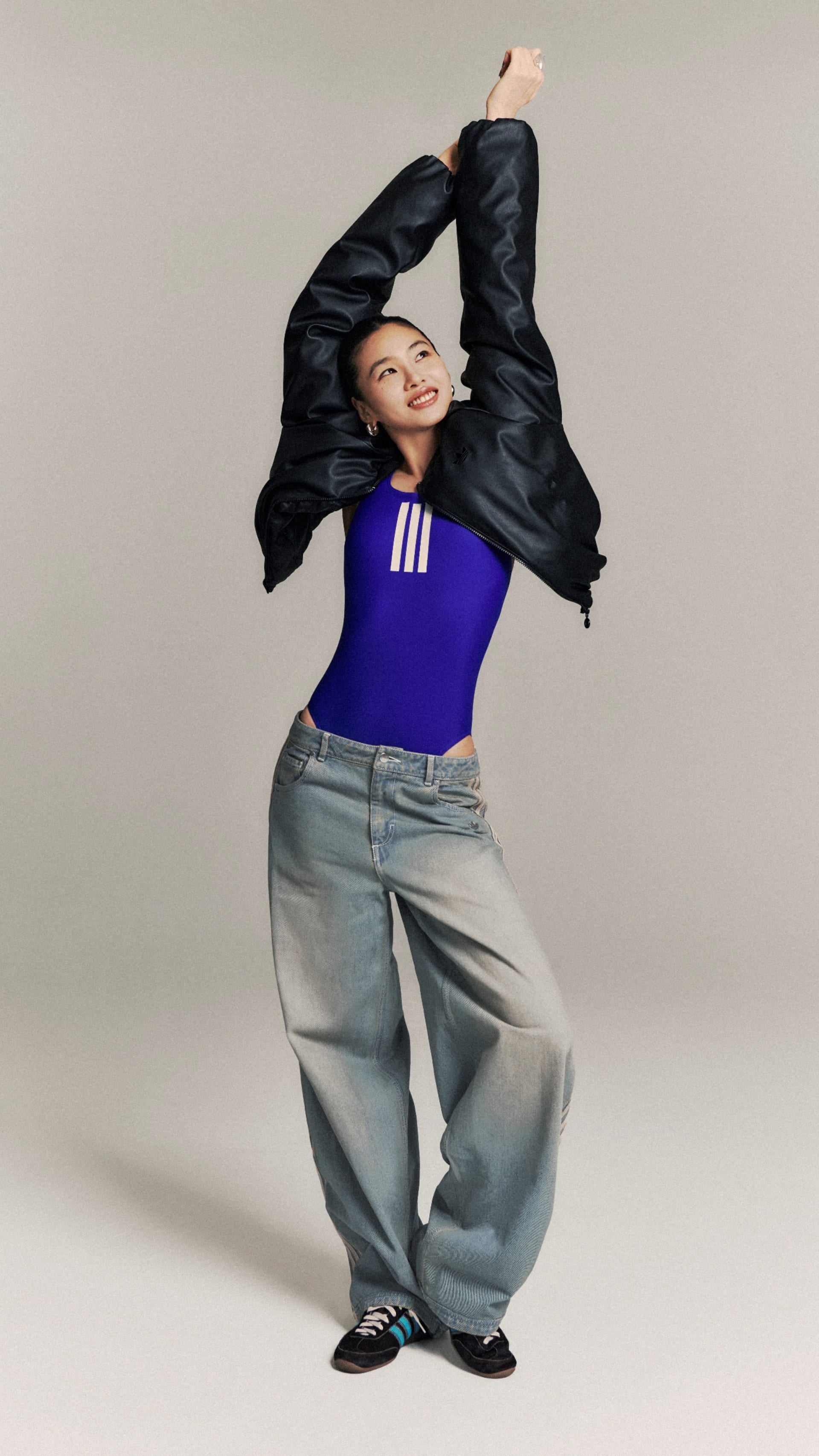 A person wearing a blue top and gray pants is striking a dynamic pose, with their arms raised and a playful expression on their face, against a plain gray background.