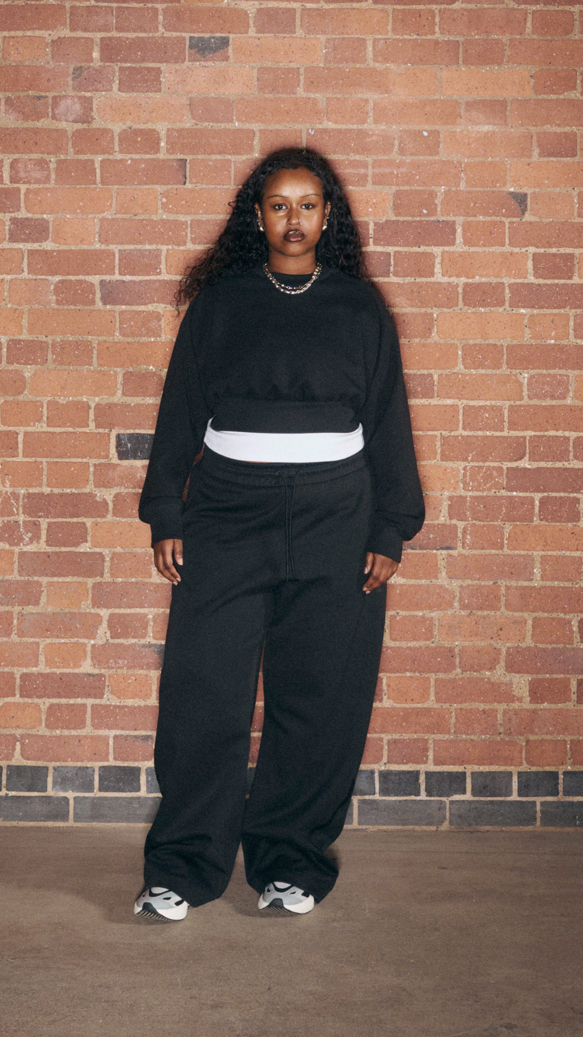 A person wearing a black hooded sweatshirt and loose-fitting pants stands in front of a brick wall.