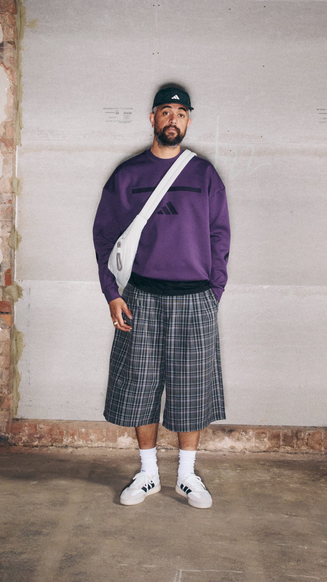 A person wearing a purple sweatshirt, plaid shorts, and white sneakers stands in front of a plain white wall.