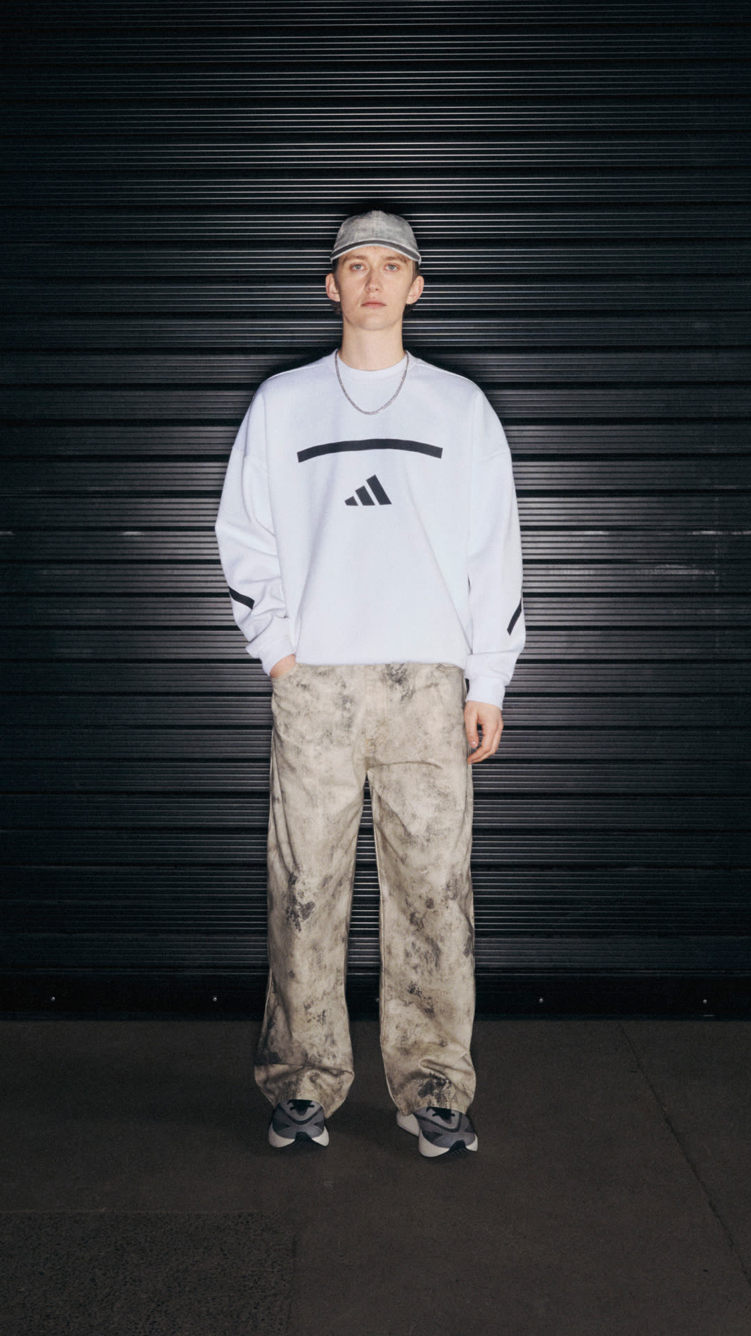 A person wearing a white Adidas sweatshirt and camouflage pants stands in front of a black metal shutter backdrop.