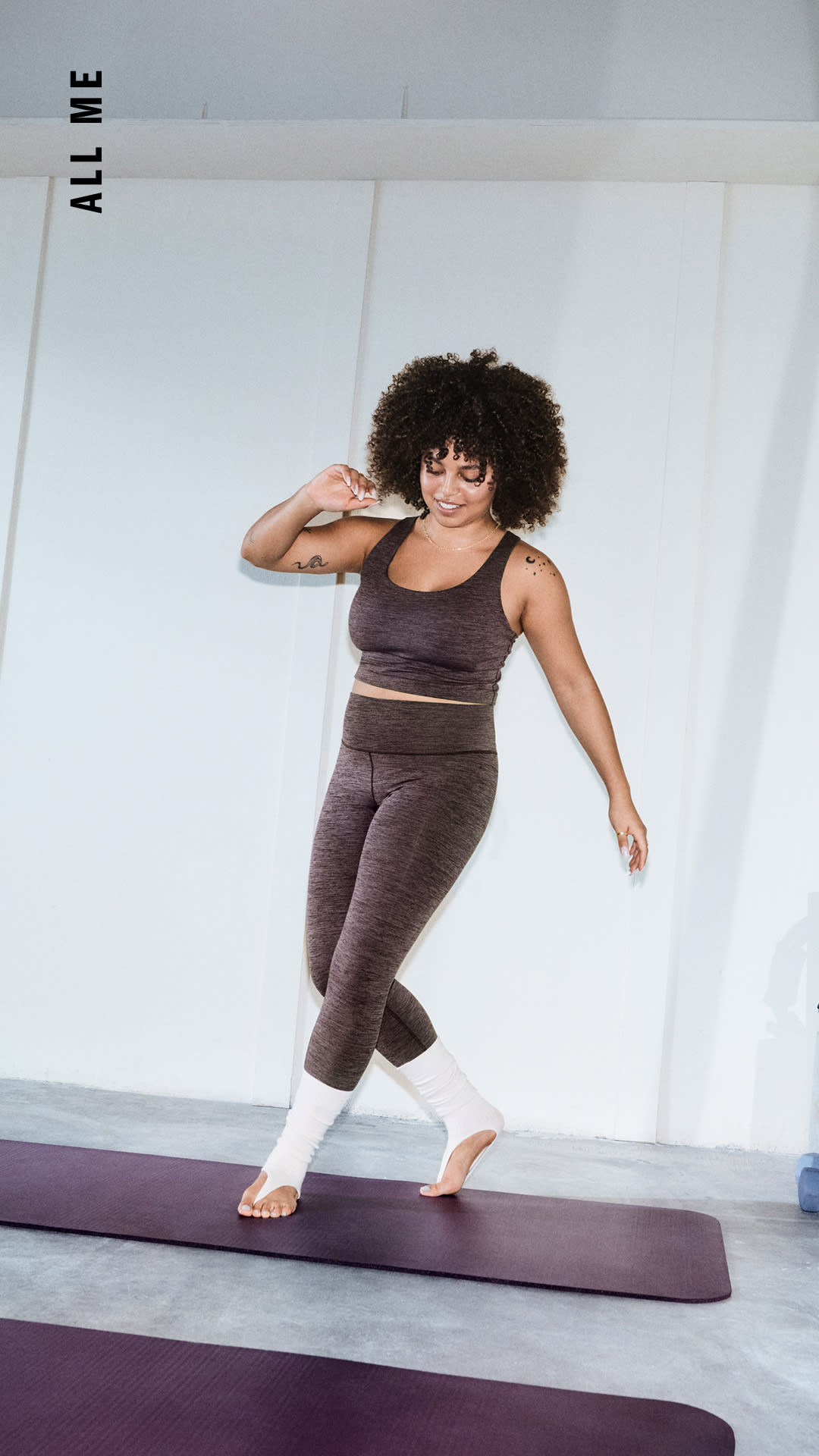 Eine Frau mit lockigem Haar steht in einem hellen, minimalistischen Raum auf einer Yogamatte.