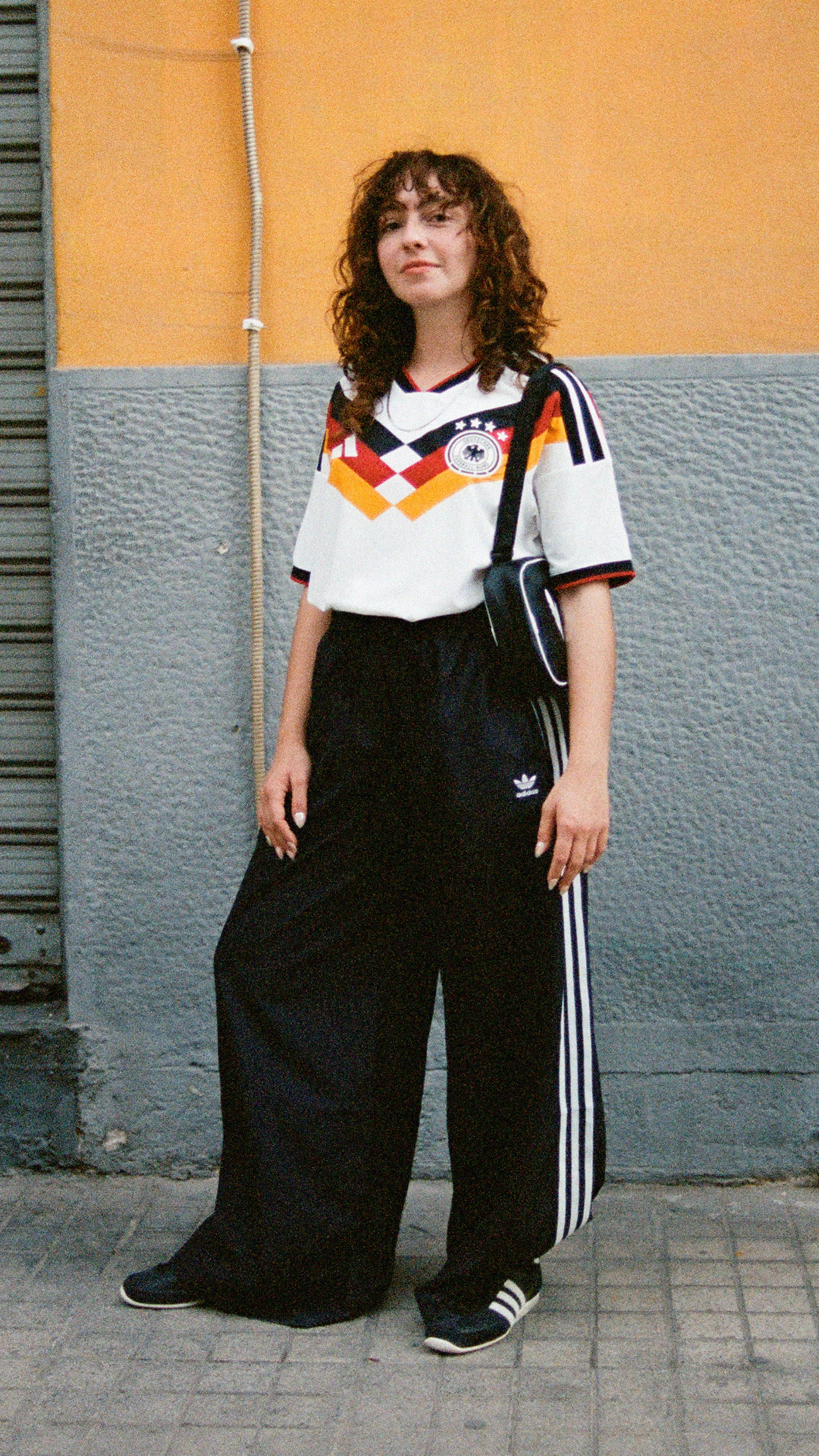 The image shows a person wearing a white and black jersey with a German flag design, standing in front of a yellow and gray wall.