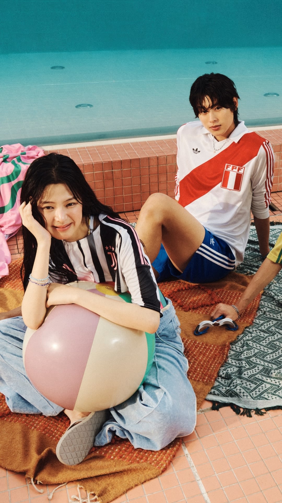 Two young people, a man and a woman, are sitting on a colorful patterned blanket by the ocean, with a bright blue sky and calm waters in the background.