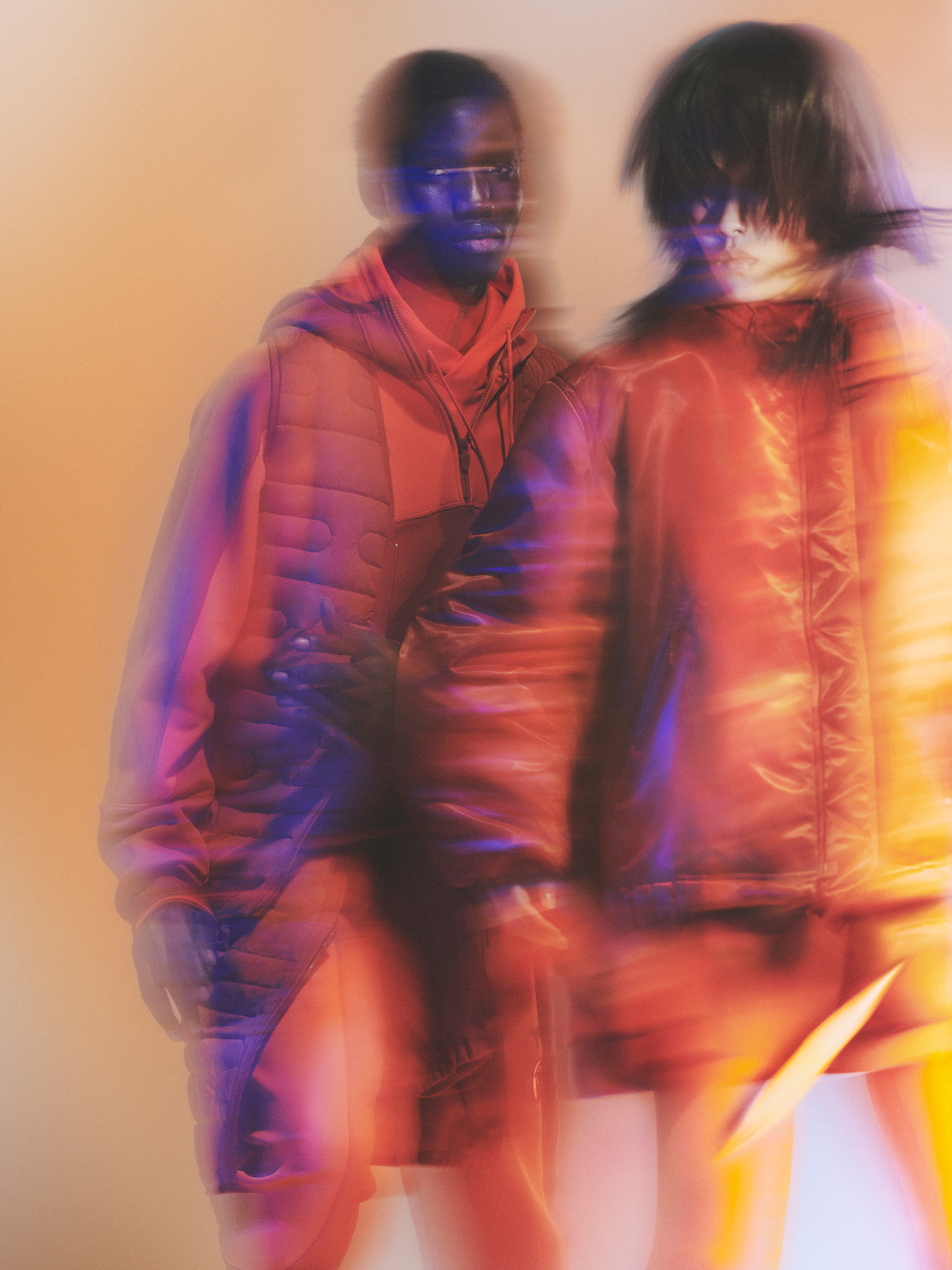 Two people wearing bright orange Y-3 jackets and hoodies look stern while gazing into the camera in front of a pale orange backdrop.
