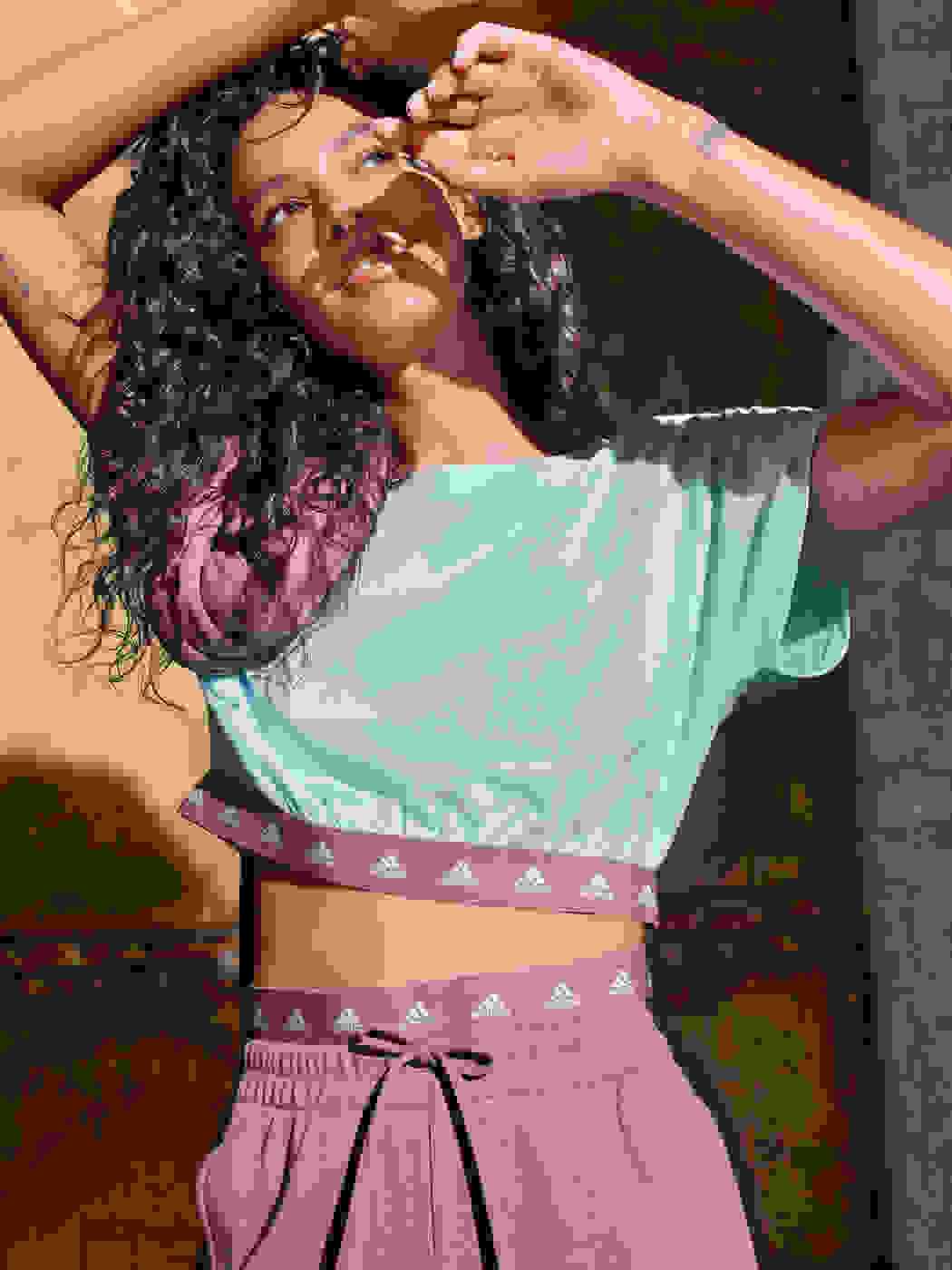 Image of female talent (brown hair) wearing pink pants and blue top, posing, one arm over the head, one hand touching the face.