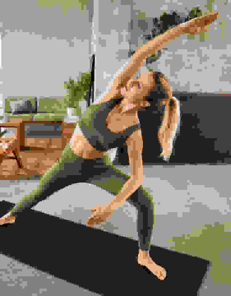A woman in an olive green bra and tights stretches on a mat in her living room.