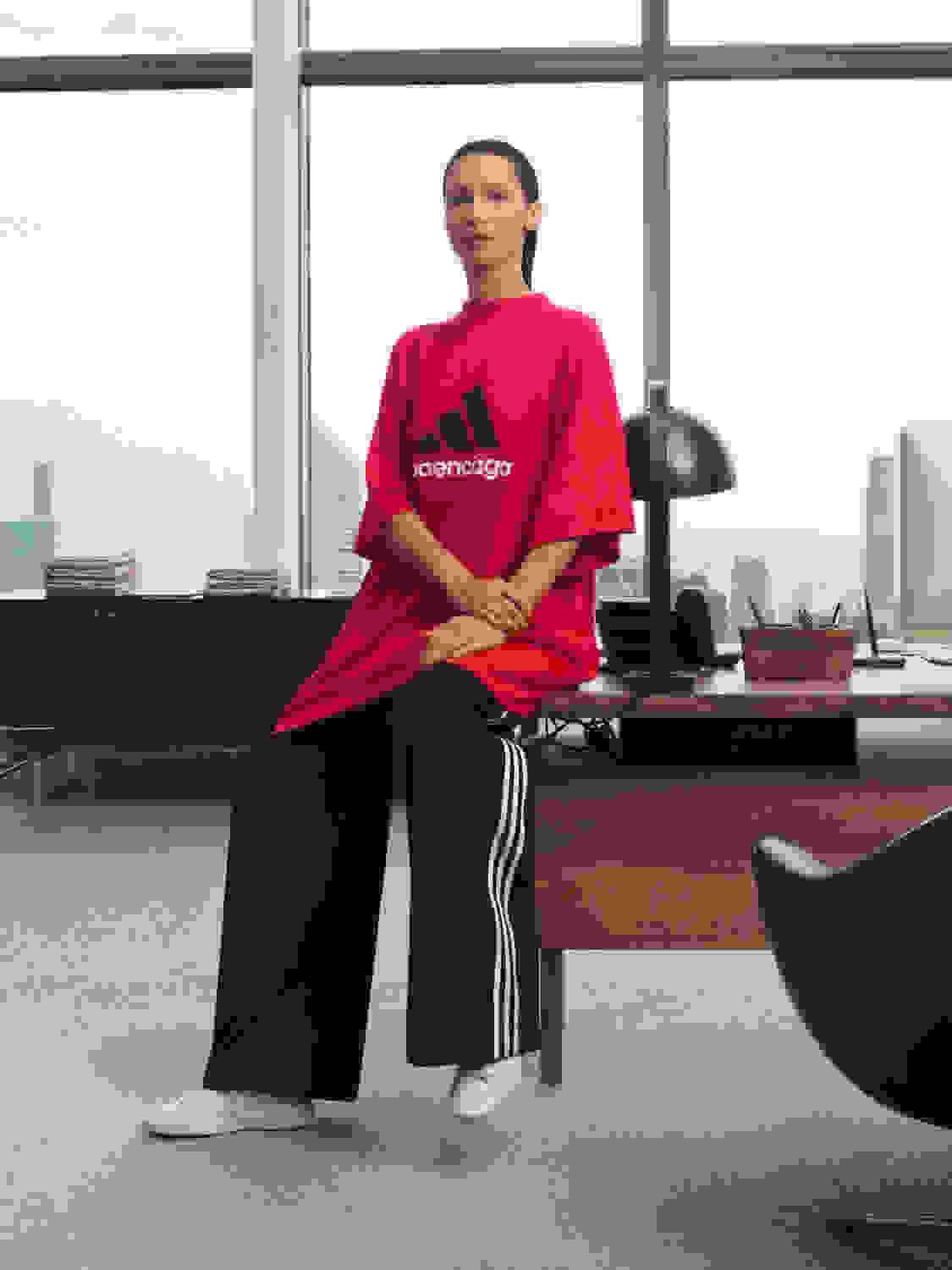 Image of Bella Hadid sitting on an office desk, wearing a red oversized shirt, black trackpants and the Stan Smiths from the BALENCIAGA / ADIDAS collection.