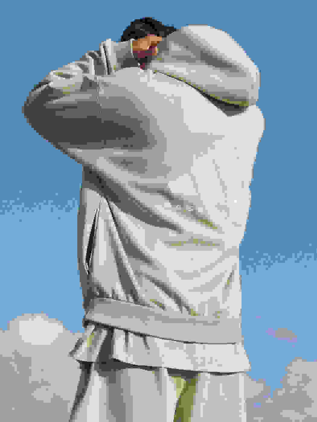 A rear view of a man standing under a blue sky, adjusting the hood of his pale green adidas sweatshirt.