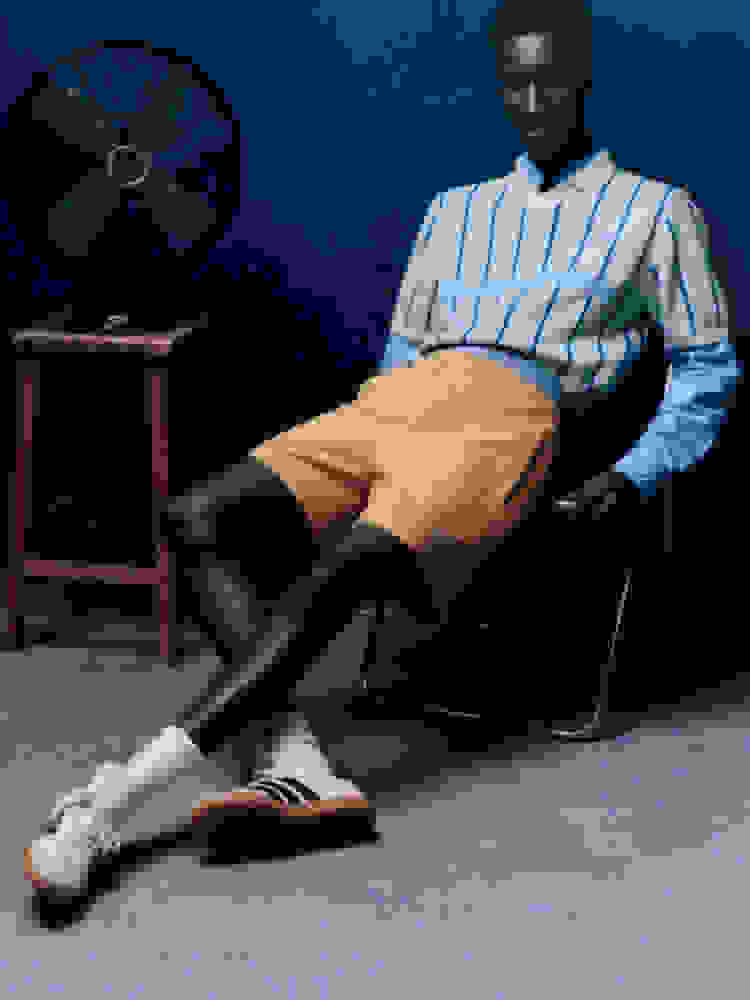 Model wearing blue shirt and orange shorts reclines on chair in dark room