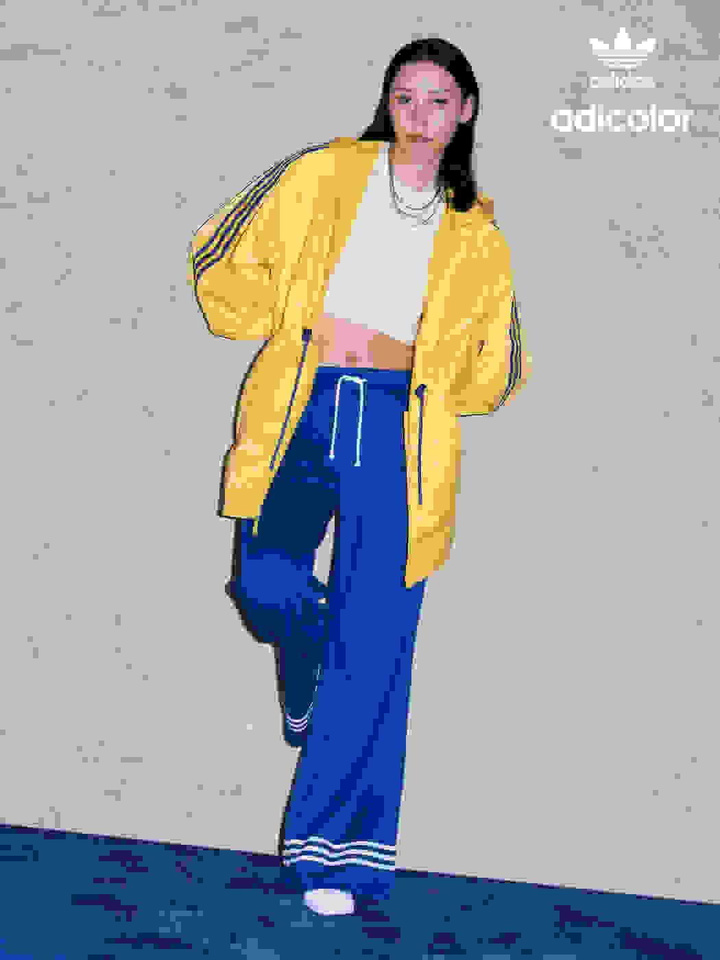 A female model wearing a white single along with a yellow jacket and blue flared trackpants from the latest adicolor collection with Gazelle shoes