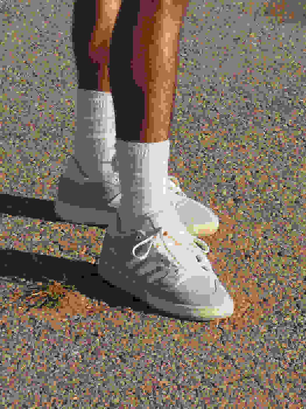 A knee-down view of a man standing on pavement, wearing white crew socks and grey adidas Forum sneakers.