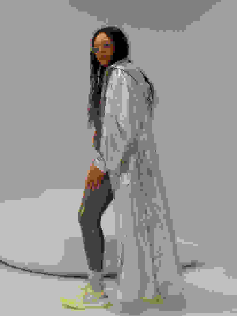 Model in metallic cape, green pants and sunglasses stands in softly-lit room.