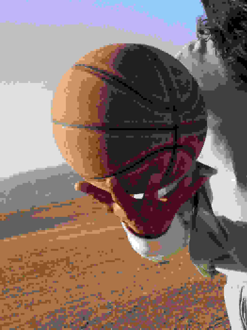 A close-up view of a right hand holding a basketball from underneath, with a dusty landscape and mountains in the background.