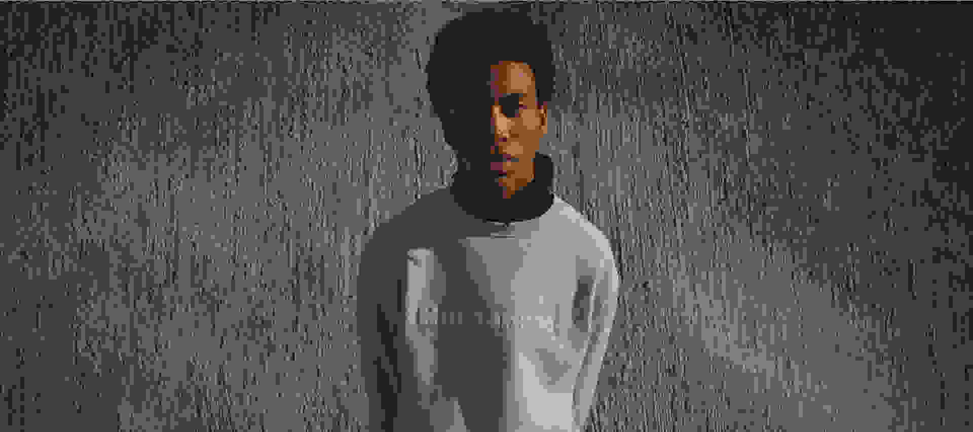 Model looks to camera wearing gray jumper against concrete background.