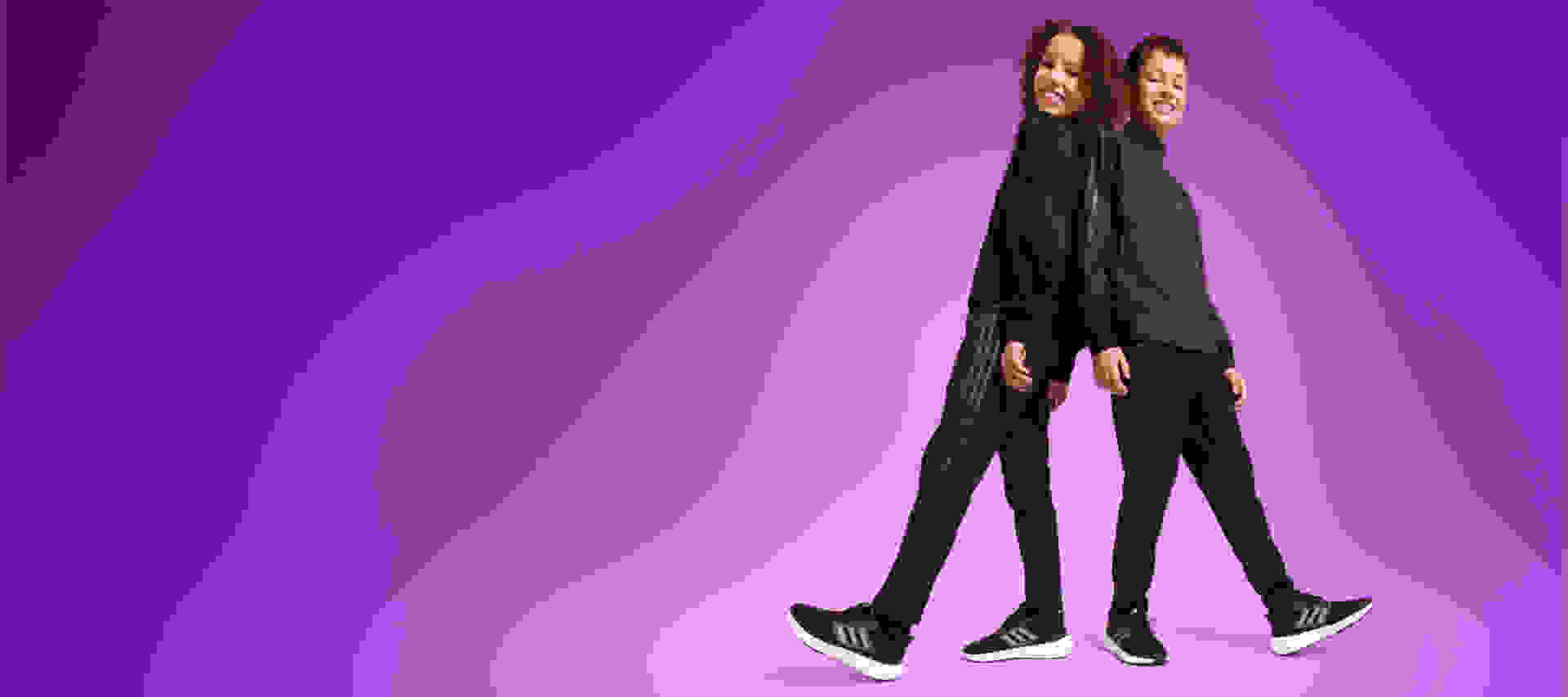 Two children, one male one female, stand smiling back to back in black adidas tracksuits and sneakers.
