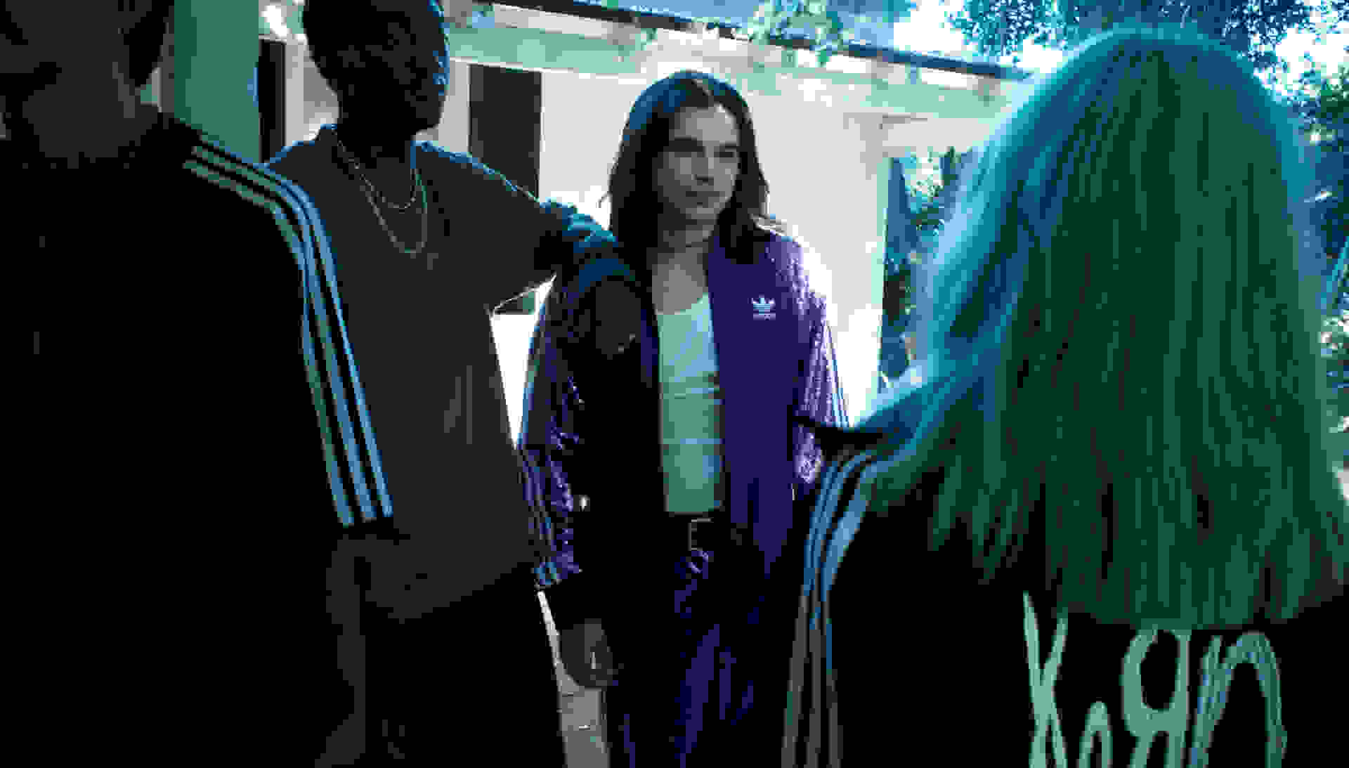A group of young people wearing the adidas Originals x KoRn collection stand in the shade in front of a house with white walls.