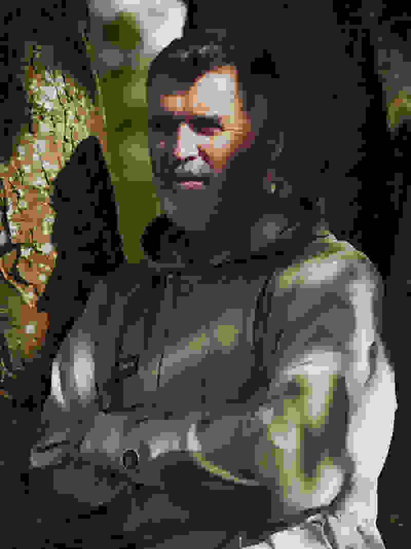 Roy Keane leans on a tree with crossed arms and a slight smile, hiding from the sun in a green and gray Spezial jacket.